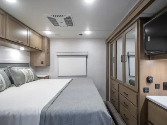Inside an RV rental bedroom: bed, overhead cabinets, AC, TV, and mirrored wardrobe for your comfort.. Thor ACE 2026