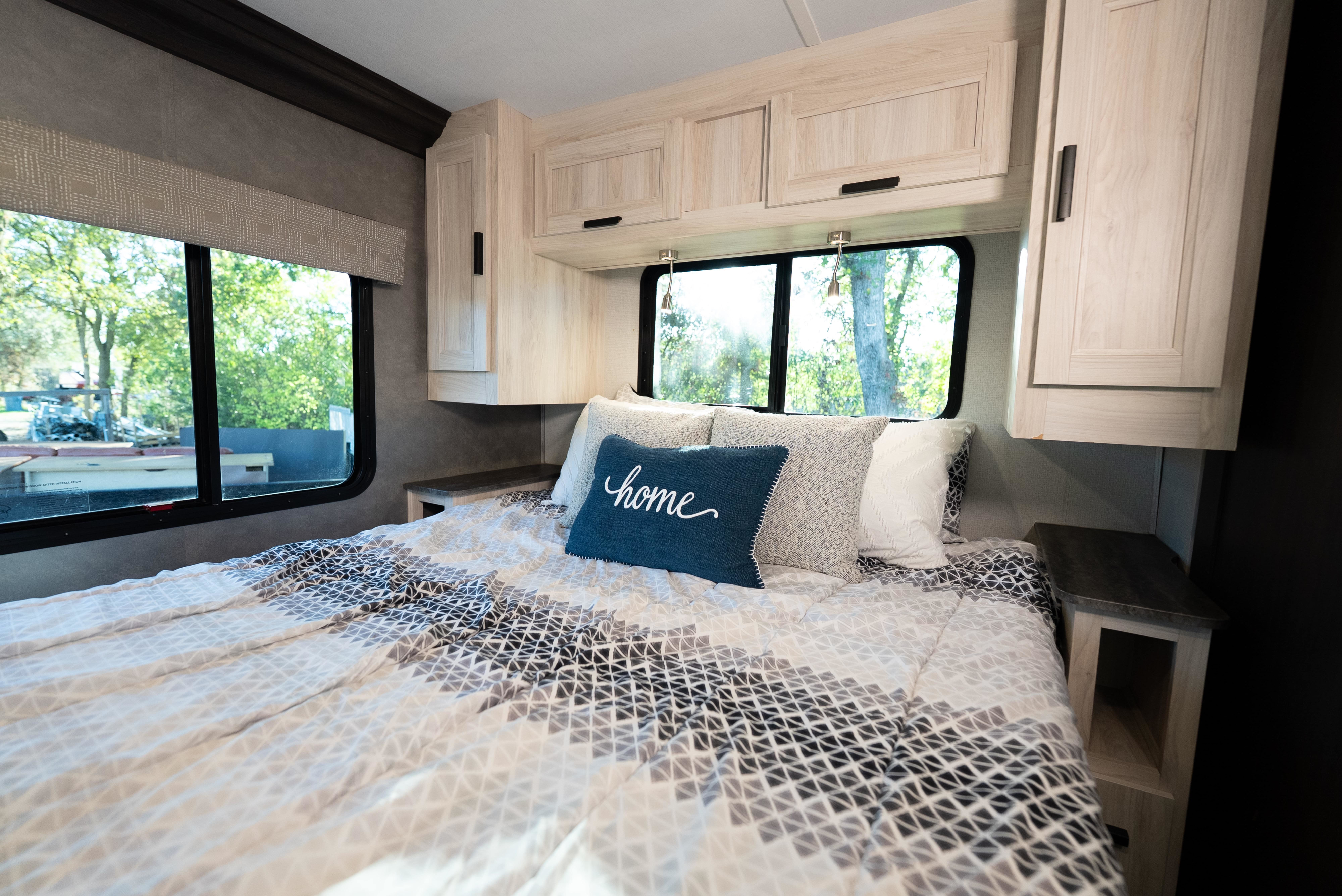 Comfortable RV rental bedroom featuring patterned bedding, "home" pillow, overhead cabinets, and bright windows.. Forest River Sunseeker LE 2022