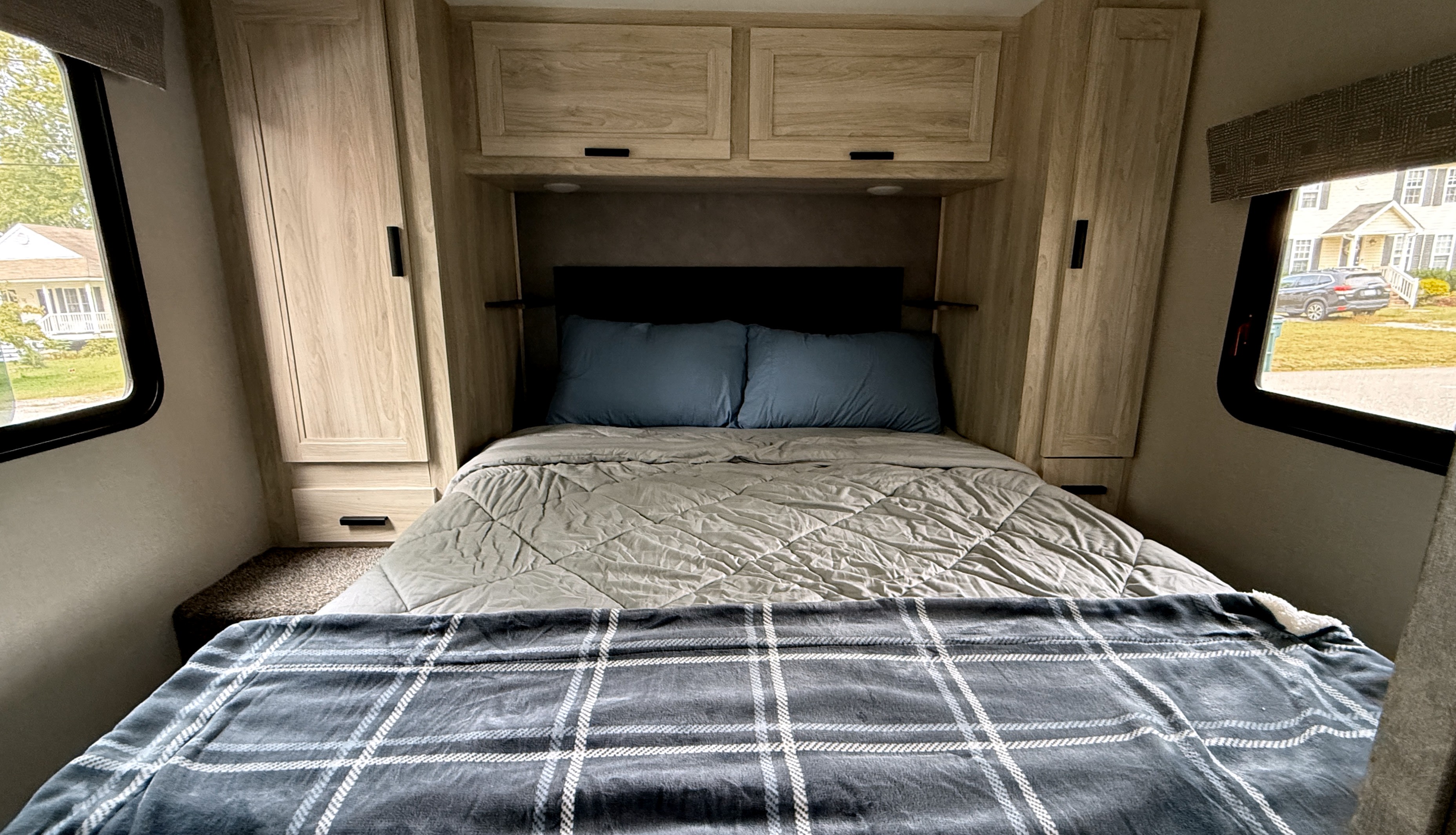 Cozy RV rental bedroom featuring a bed with blue pillows, a plaid blanket, and light wood cabinets. Windows offer an outside view.. Forest River Sunseeker LE 2023