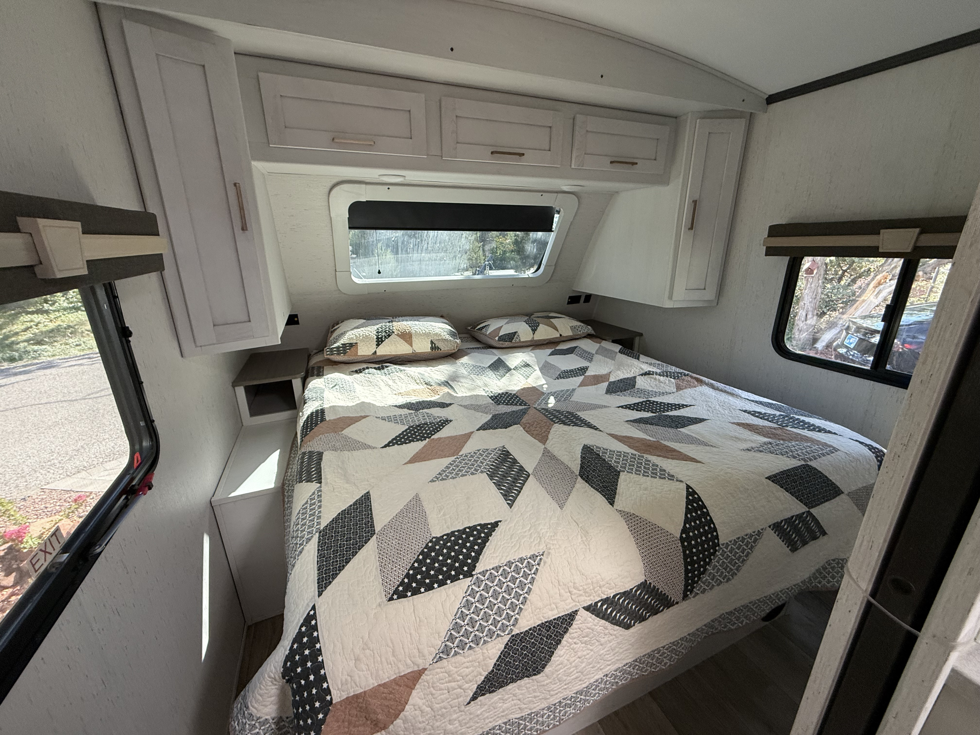 Experience comfort in this RV rental bedroom: a cozy bed with a geometric quilt, overhead cabinets, and bright windows.. Astoria Astoria Trailer 2022