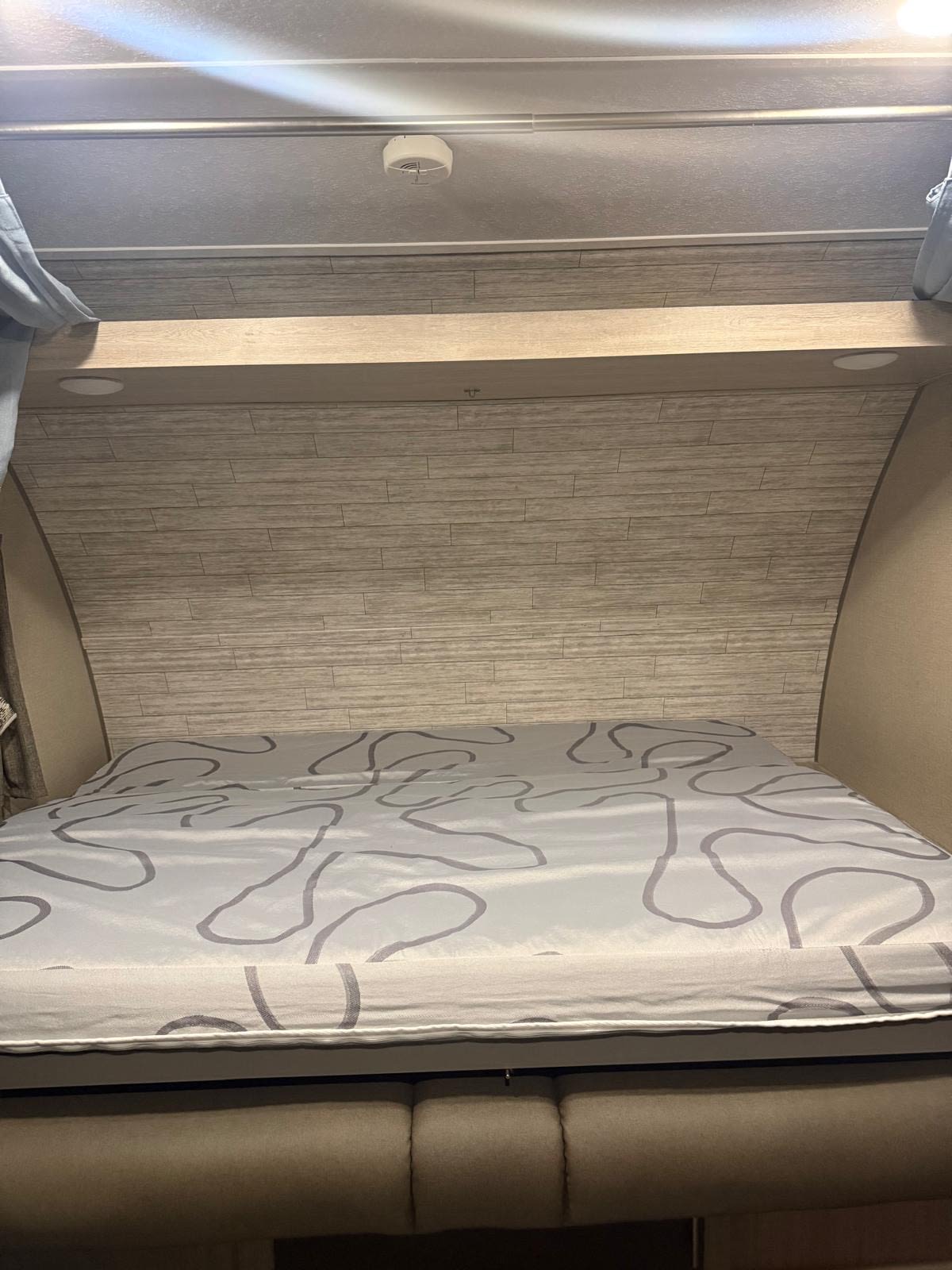RV rental bed with patterned sheet, wood-look headboard, shelf lighting, and ceiling-mounted smoke detector.. Forest River Aurora Aurora 2023