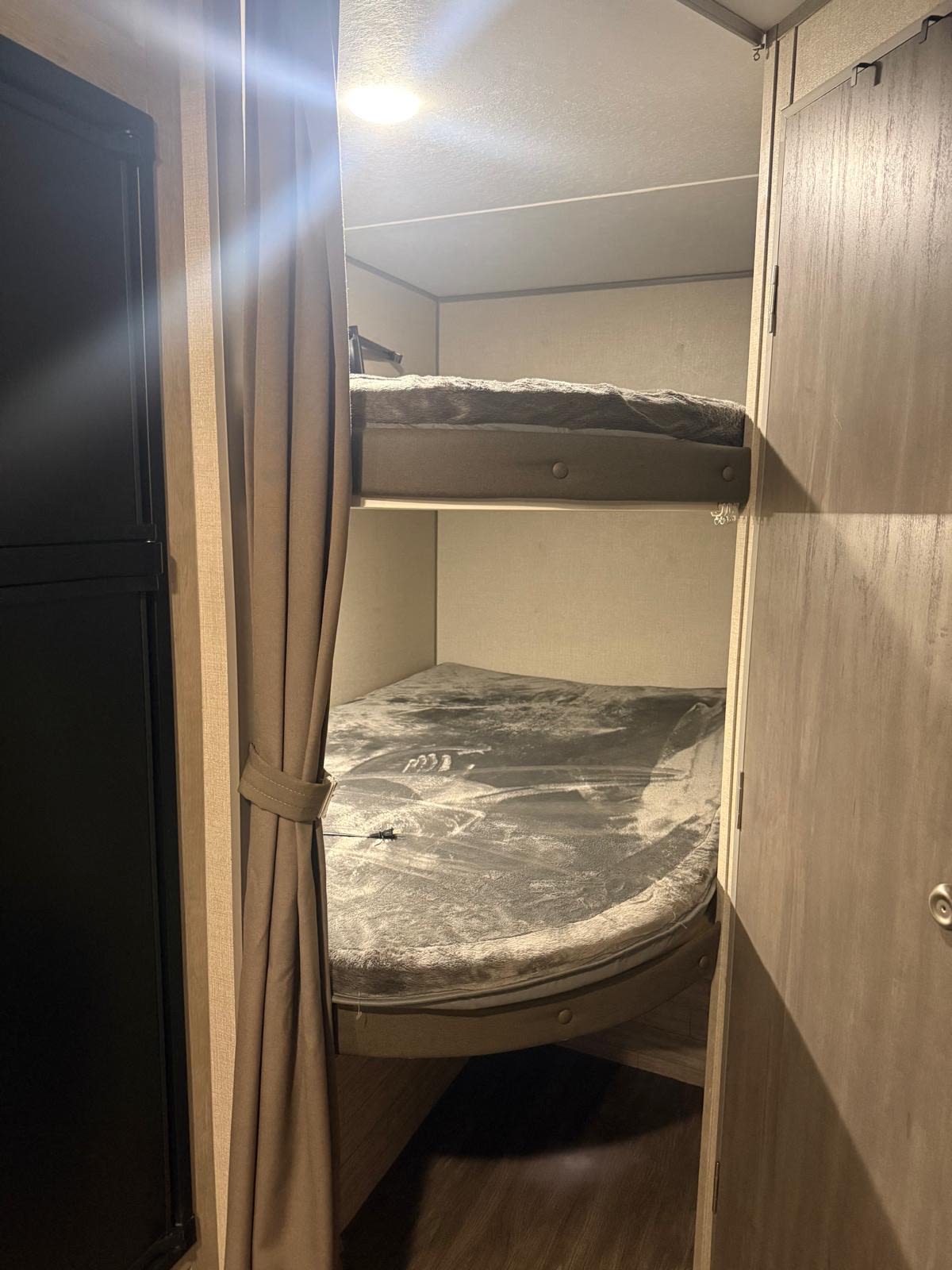 This rv rental features two bunk beds with grey bedding in a compact space. A tied-back curtain and dark appliance are visible left.. Forest River Aurora Aurora 2023