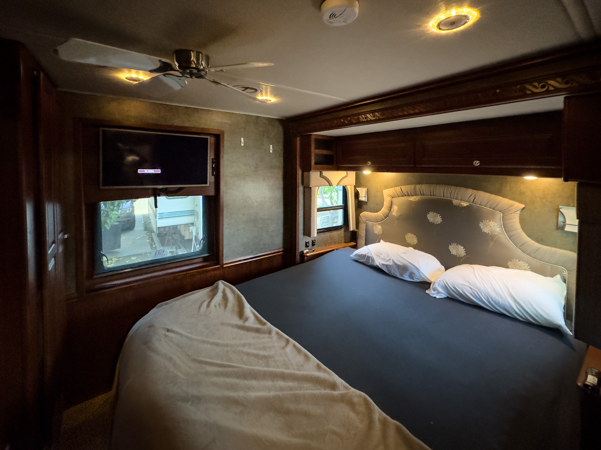 This cozy RV rental bedroom features a comfortable bed, mounted TV, ceiling fan, and warm lighting with rich wooden cabinetry, ideal for travel.. Winnebago Adventurer 2015