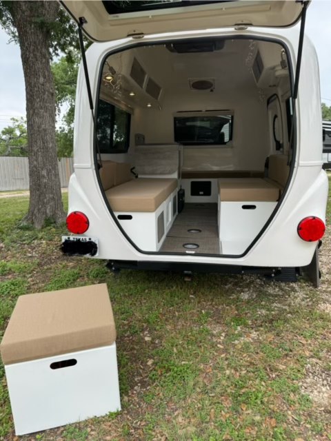 Open rear hatch of a white **rv rental**, showing a clean interior with tan benches, TV, and white cabinetry. An ottoman sits on the grass.. Happier Camper HC1 2022