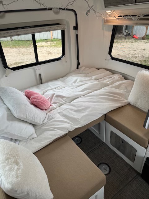 This RV rental offers a cozy sleeping area, featuring a white bed with pillows, large windows, and cushioned seating.. Happier Camper HC1 2022