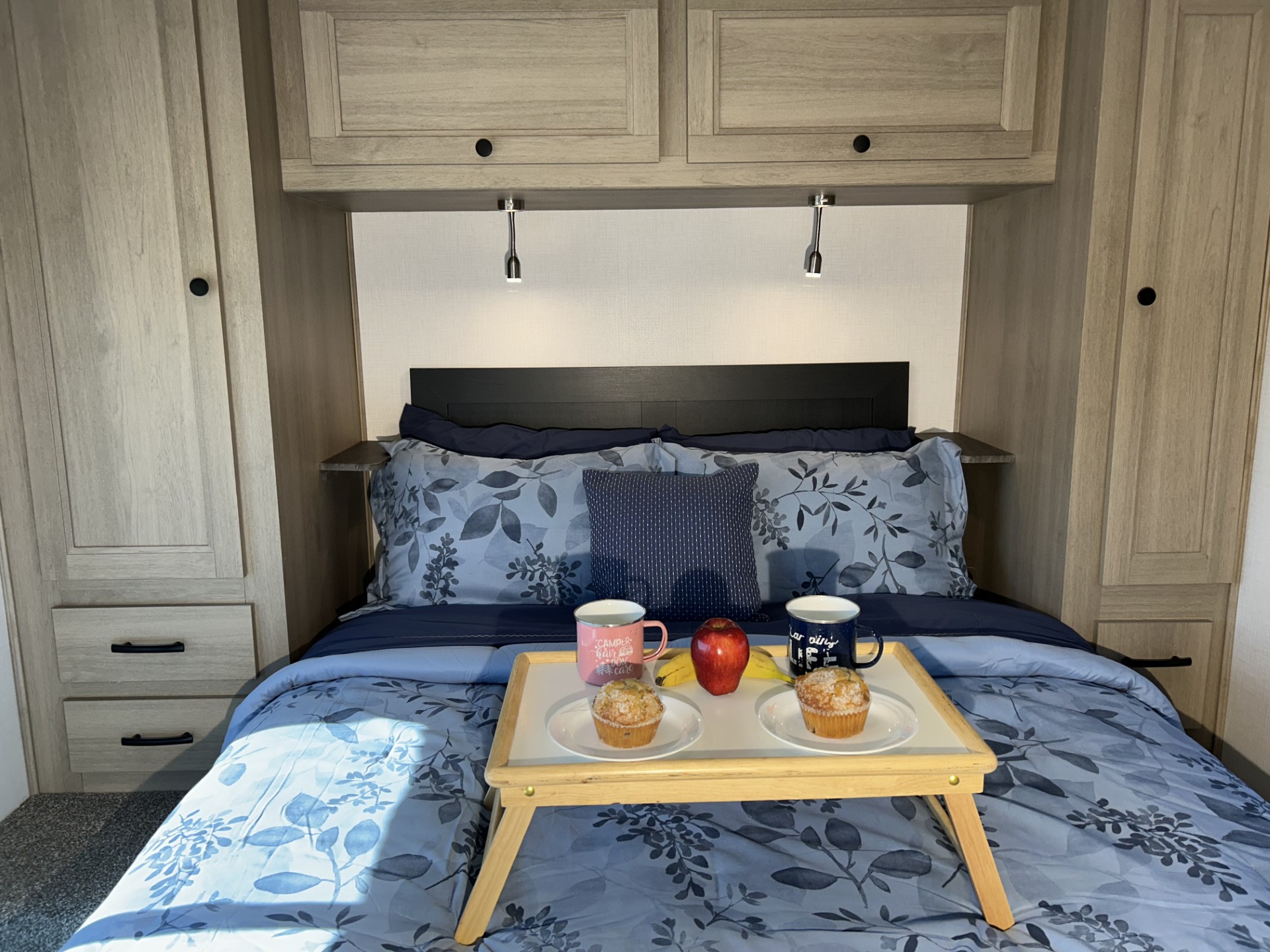 RV rental bedroom with breakfast in bed: muffins, fruit, and mugs on blue floral bedding, plus compact storage.. Forester LE 2026