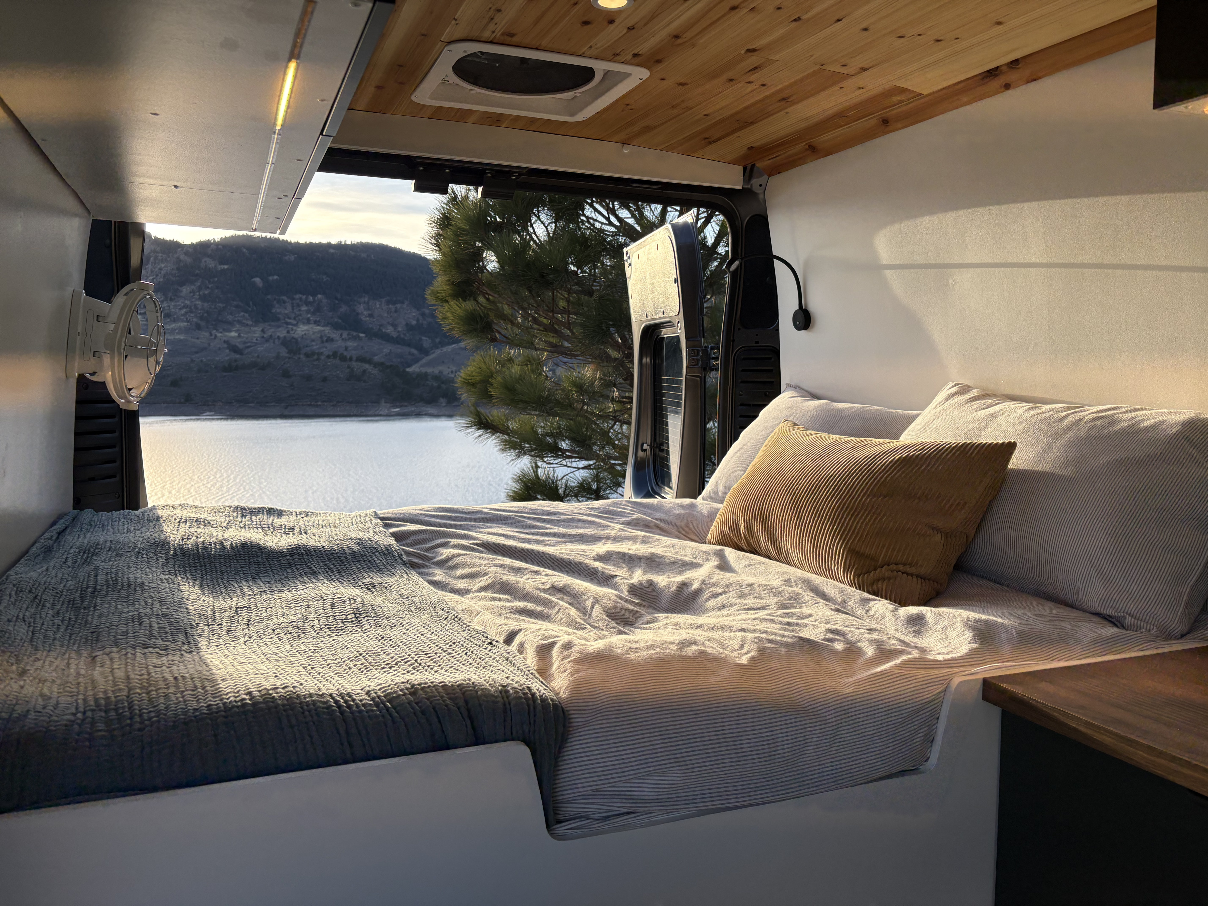 Serene RV rental interior: A comfy bed with a view of a tranquil lake and pine tree through the open van doors.. Ram Promaster 2019