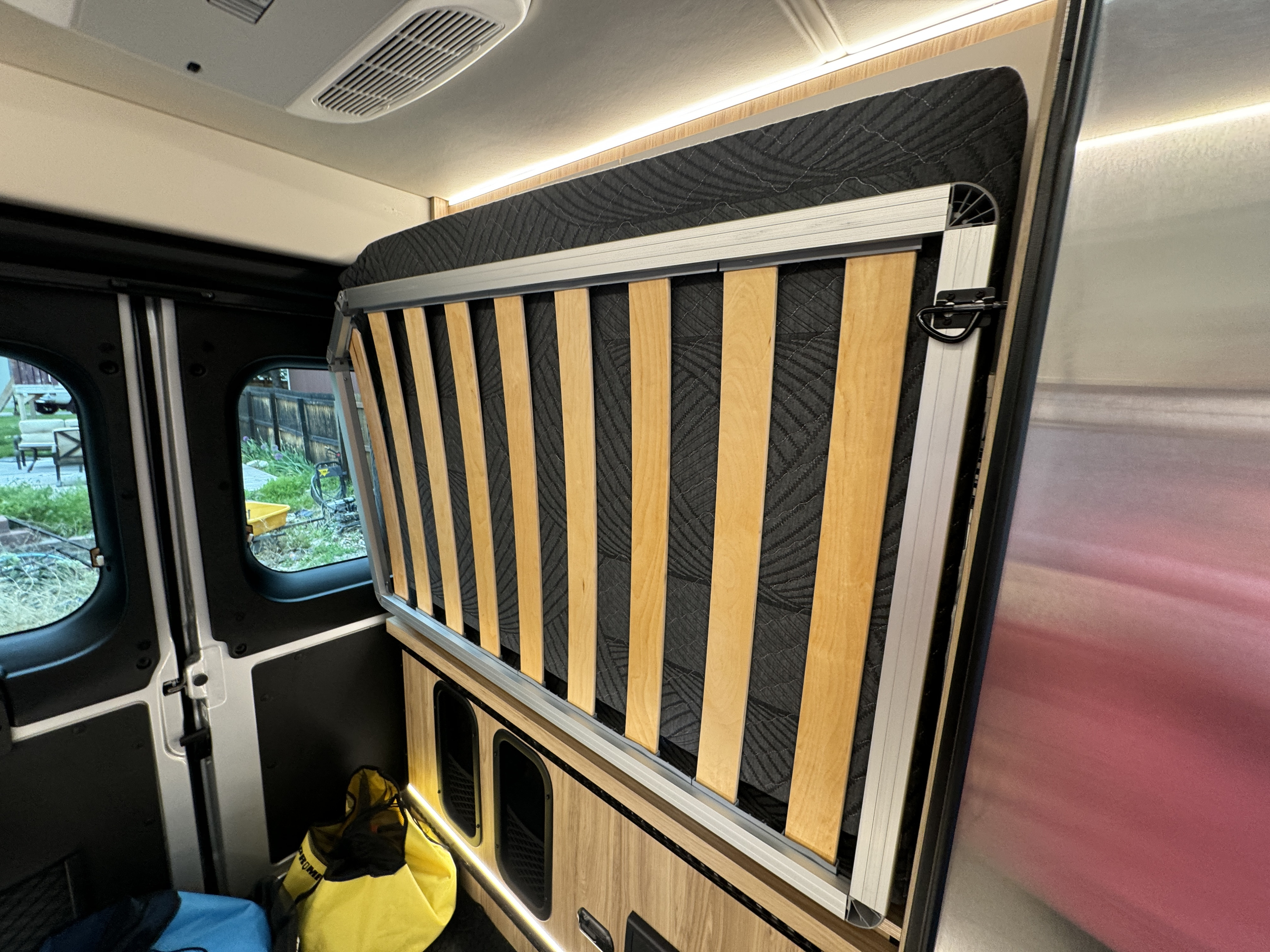Inside an RV rental, a fold-up bed with wooden slats is visible against a quilted wall. LED lights illuminate the modern camper van interior and storage.. Forest River Coachmen Pixel 2026