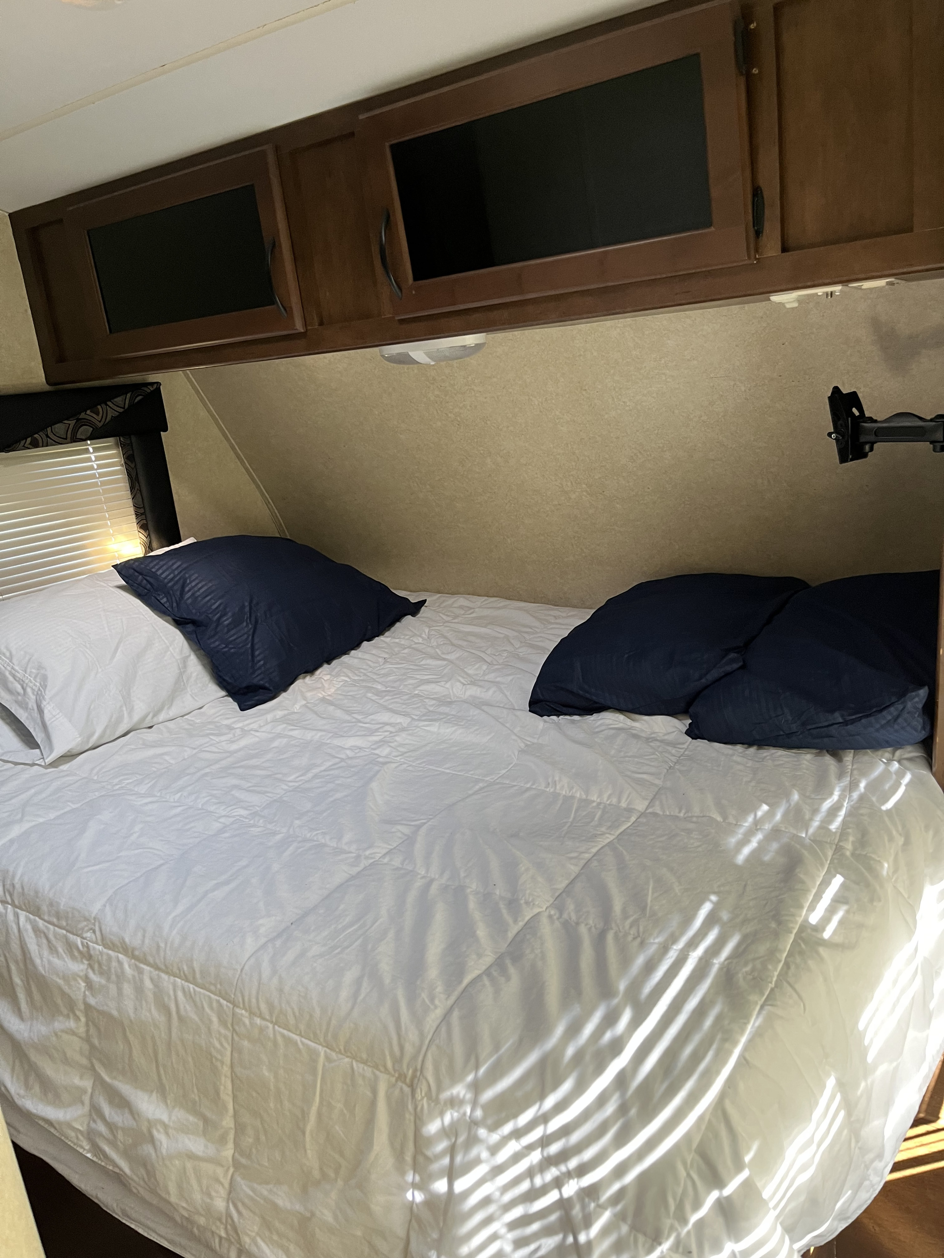 Inside an RV rental: a cozy bedroom with a white comforter, navy pillows, overhead cabinets, and window blinds.. Pacific Coachworks Sandsport 2015