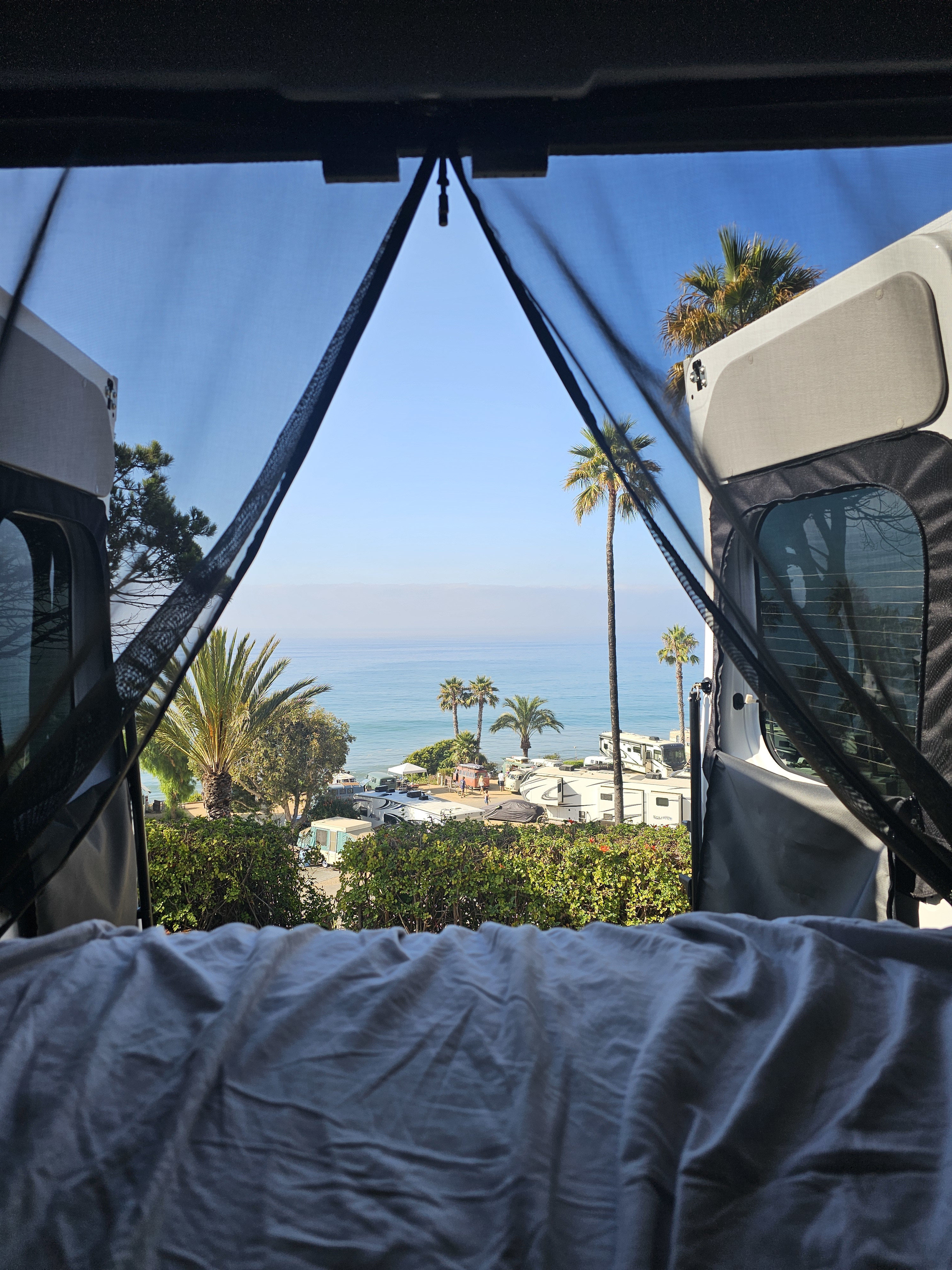 Ocean view from an RV rental, looking through open doors & bug screen at palm trees & a lively beachside campground.. Winnebago Solis 59PX 2025