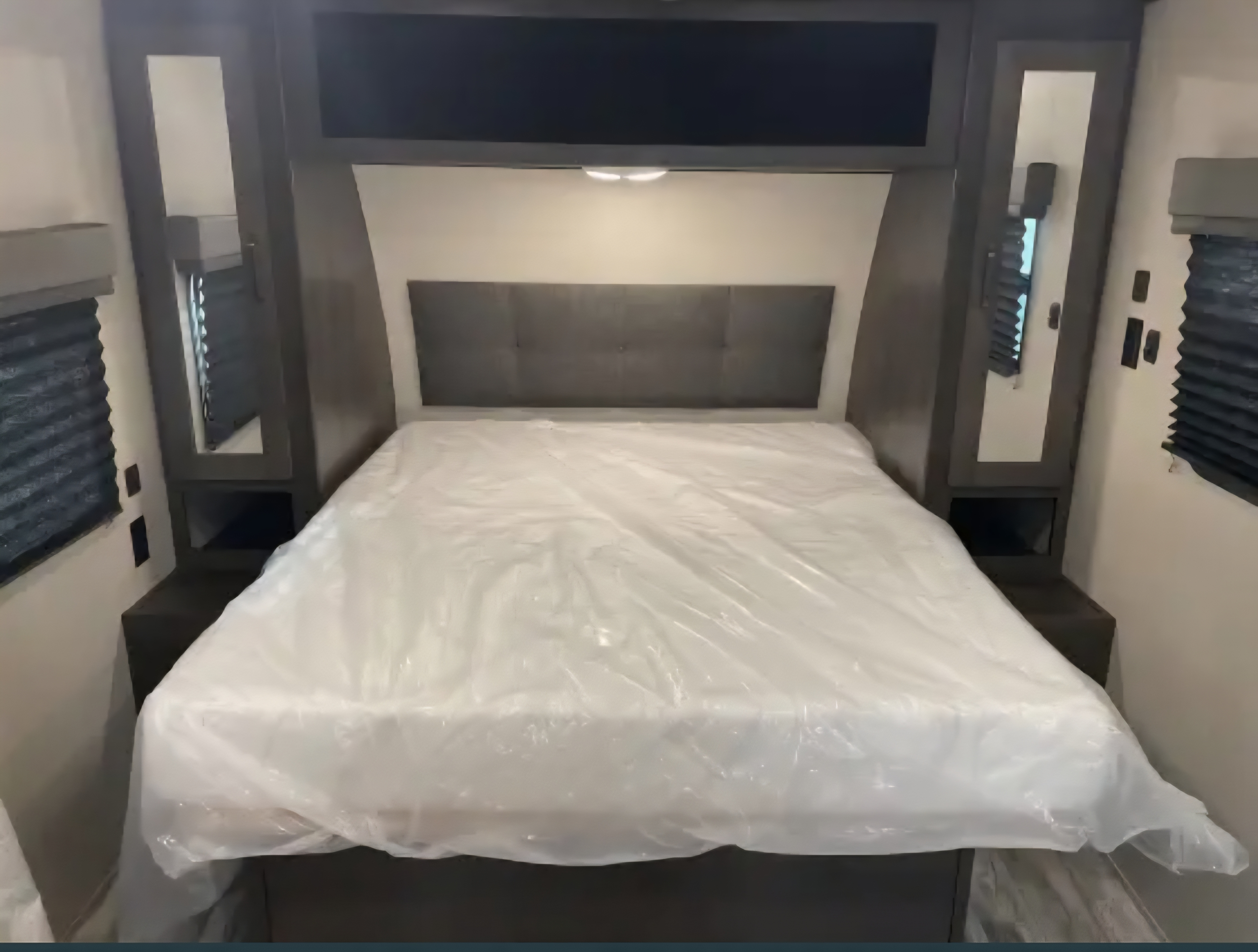 This RV rental bedroom features a clean bed with protective plastic, an upholstered headboard, and built-in storage.. Grand Design Transcend Xplor 2022