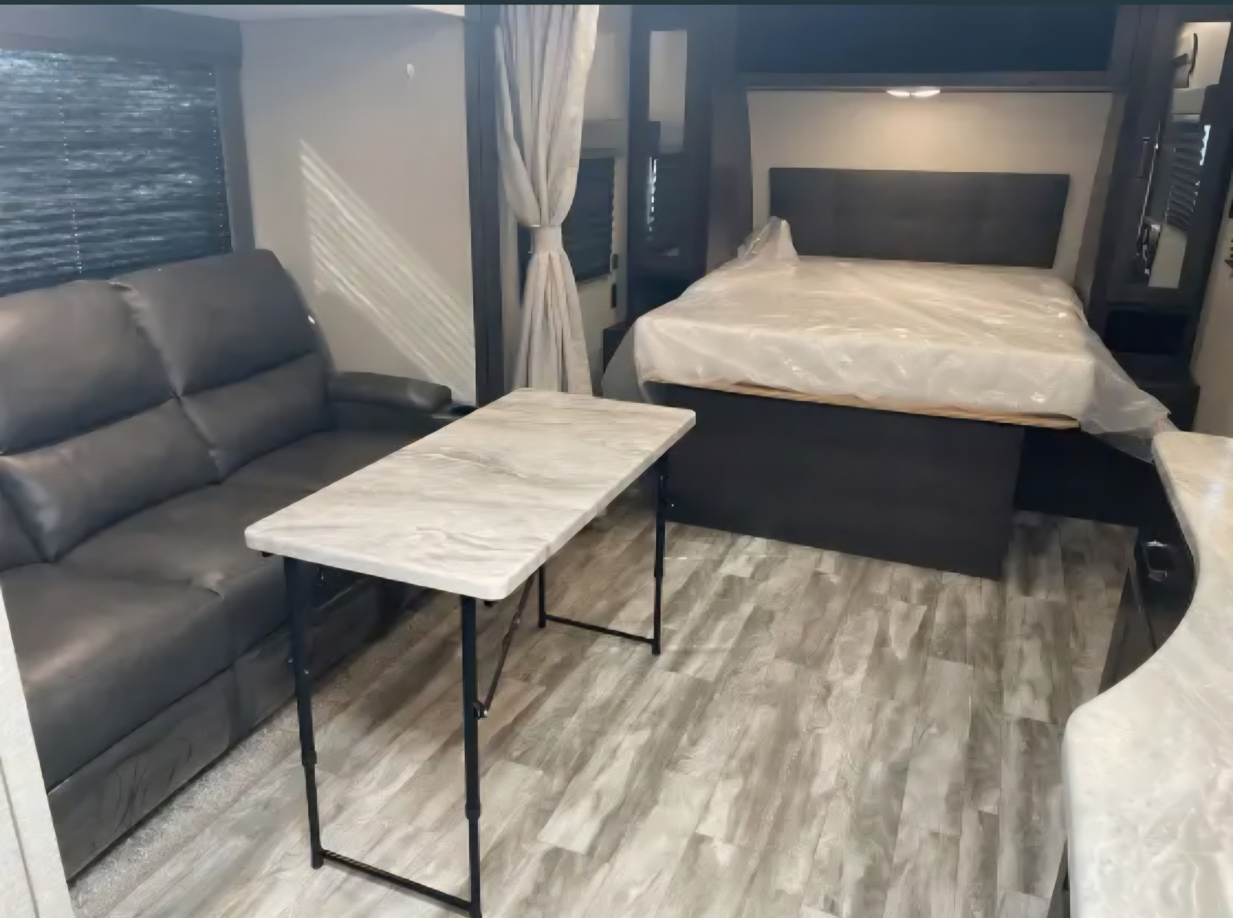 RV rental interior: a dark sofa, light folding table, and a bed with a covered mattress in a modern, compact space.. Grand Design Transcend Xplor 2022