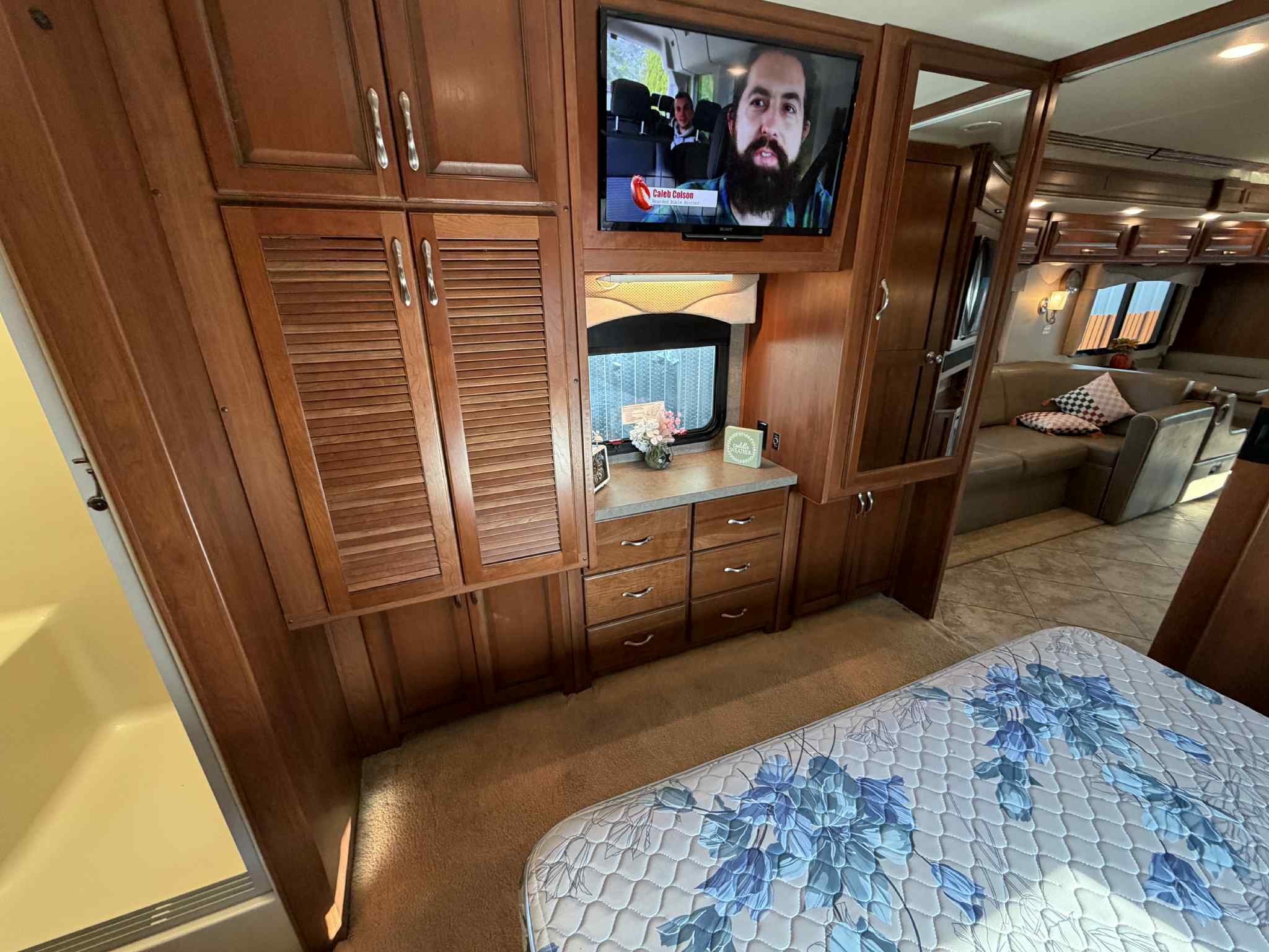 This RV rental bedroom features wood storage, a TV, window, and shower, next to a comfy patterned mattress.. Fleetwood Bounder 35k 2016