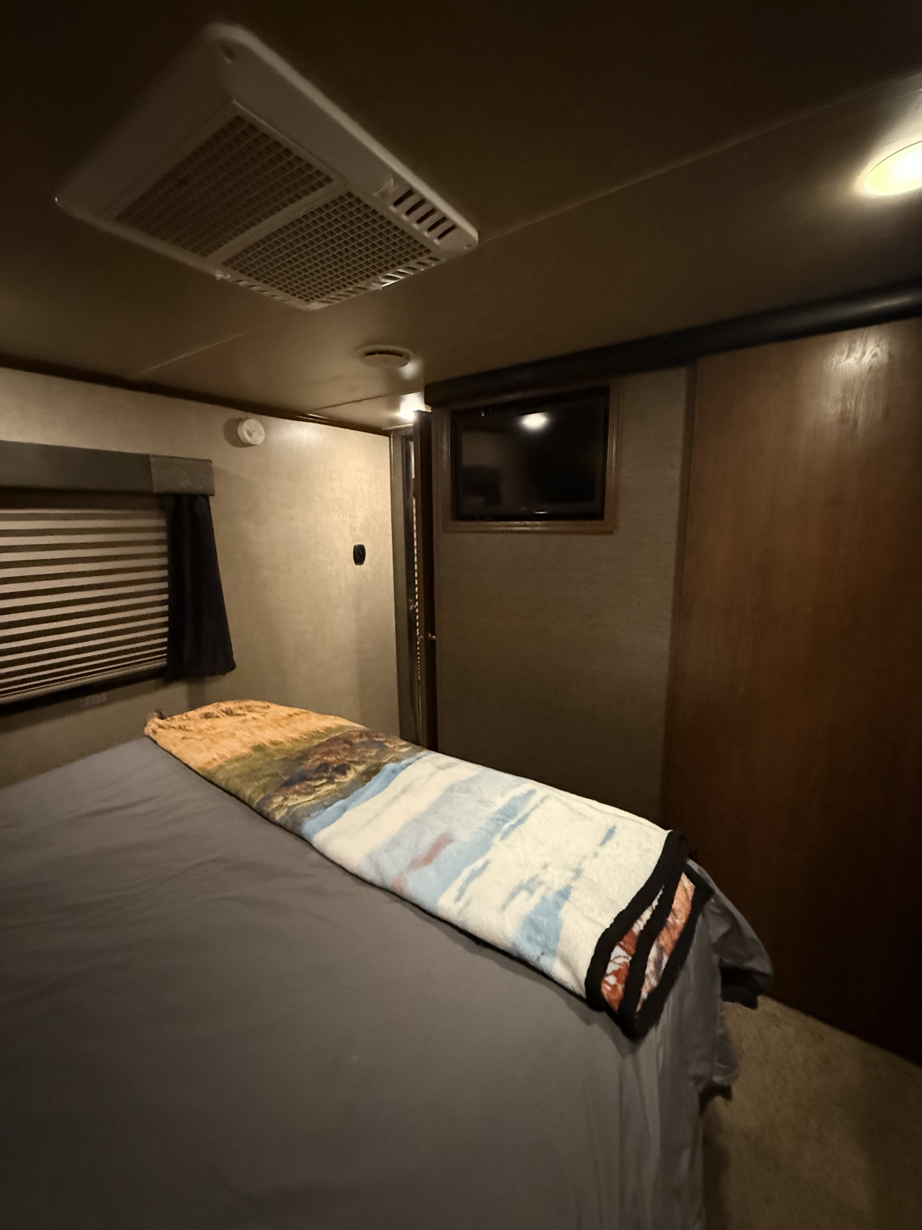 Interior of a cozy RV rental bedroom features a bed with a scenic blanket, mounted TV, AC, and window blinds.. Keystone RV Fuzion Toy Hauler 2015