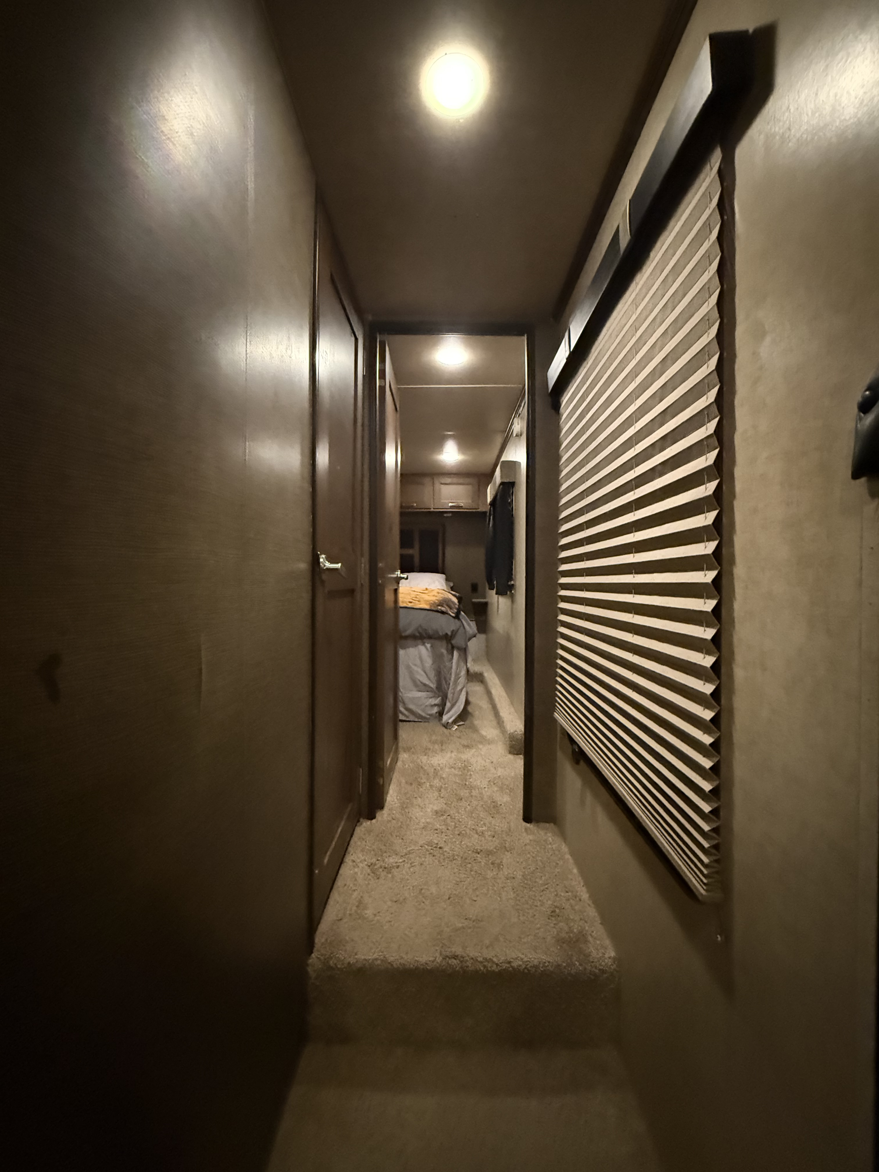 Carpeted hallway in an RV rental with a bright ceiling light, door, and pleated blind, leading down steps to a bedroom.. Keystone RV Fuzion Toy Hauler 2015