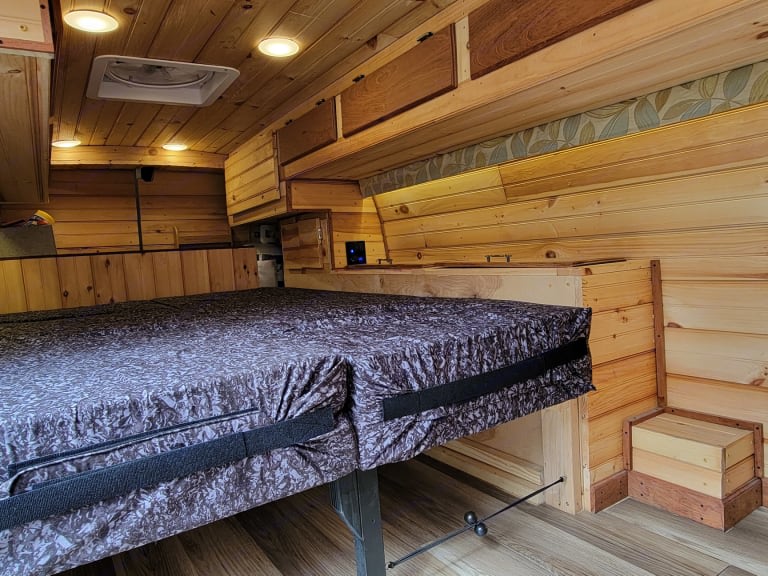 Cozy wood-lined RV rental interior with a comfy bed, overhead storage, ceiling lights, a vent fan, and wooden steps.. Gmc Savanna 2014