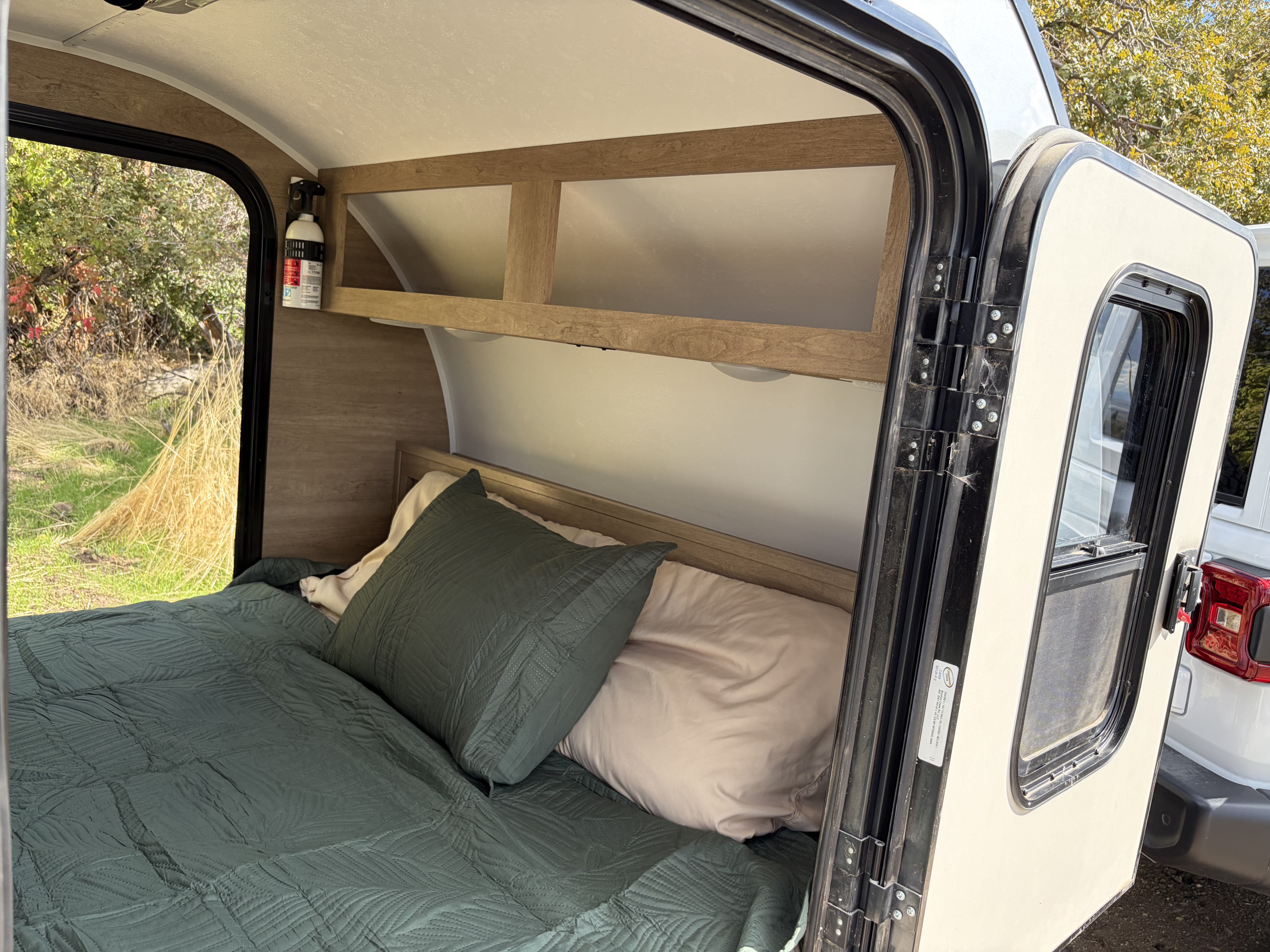 Cozy RV rental interior with a comfortable bed, overhead shelf, and a view of nature outside the open door. Ideal for camping.. Braxton Creek Braxton Creek Trailer 2020