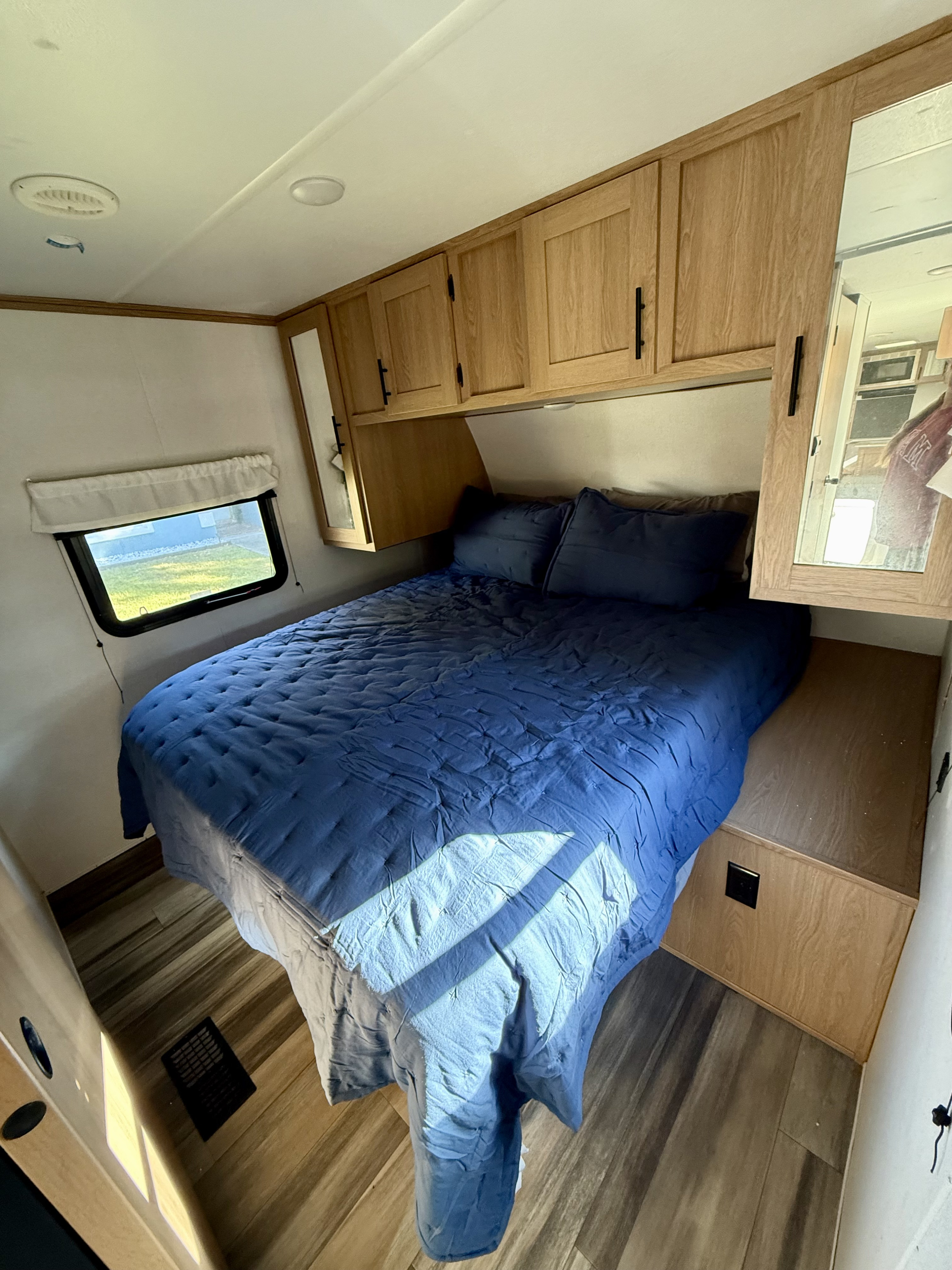 Inside an RV rental: a bright bedroom with a blue quilted bed, overhead storage, mirrored cabinets, window, and wood-look floor.. Avenger Avenger Trailer 2022