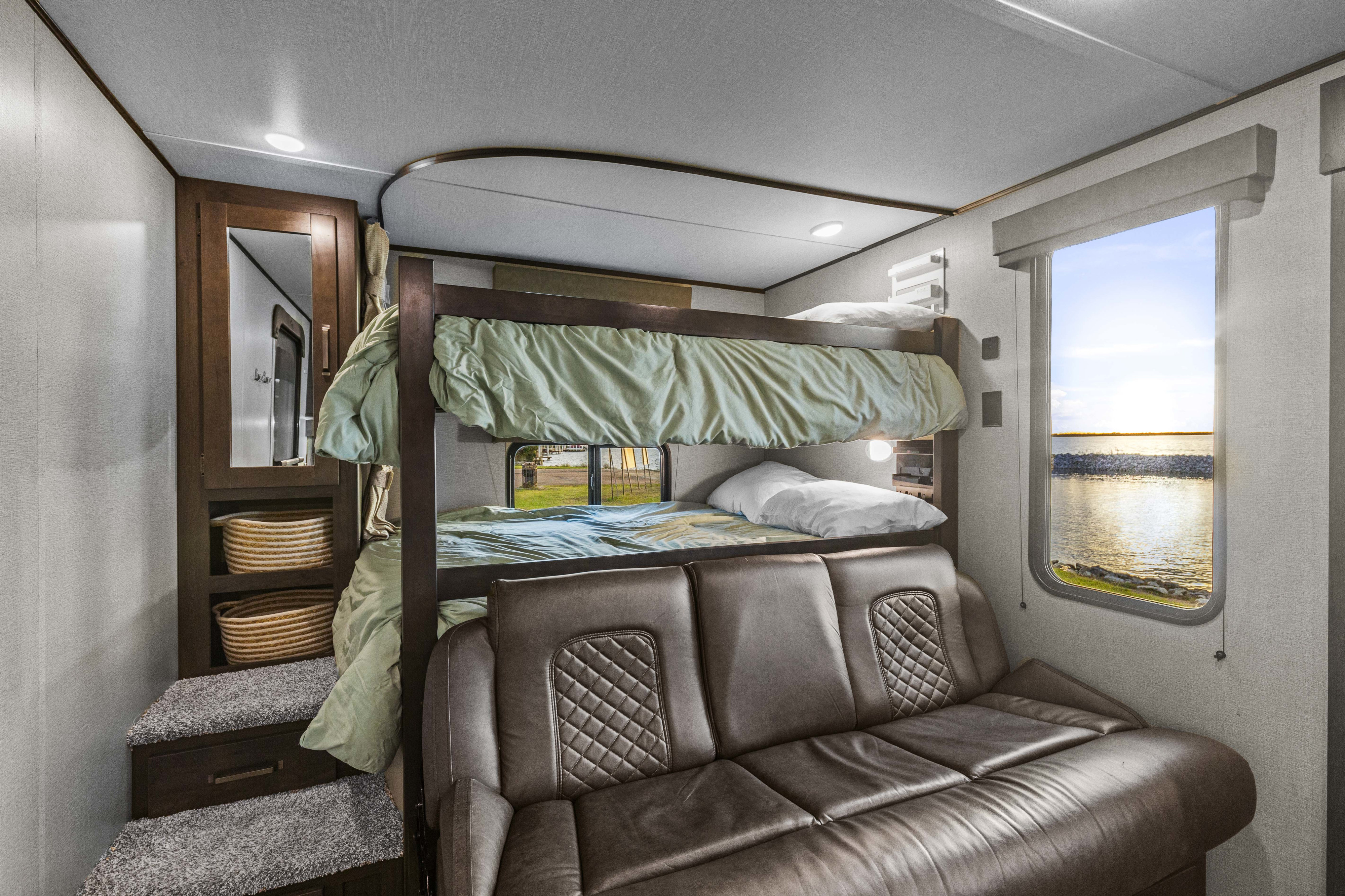 Cozy RV rental interior featuring bunk beds, a sofa, and a scenic window view of a lake at sunset.. Grand Design Transcend Xplor 2023