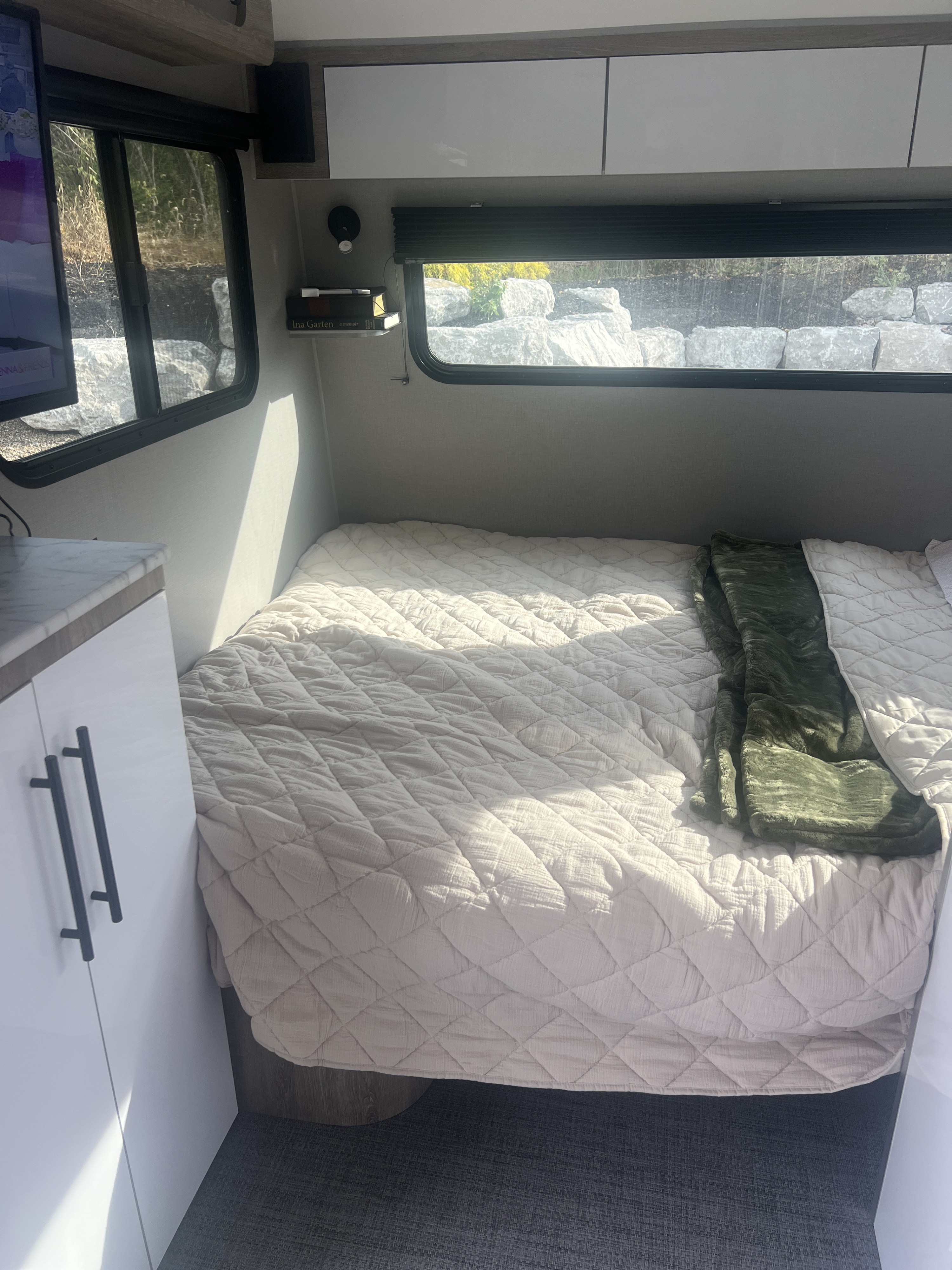 Inside a modern RV rental: a comfy bed with a quilted spread, window view of rocks, and compact storage.. InTech RV Sol 2023