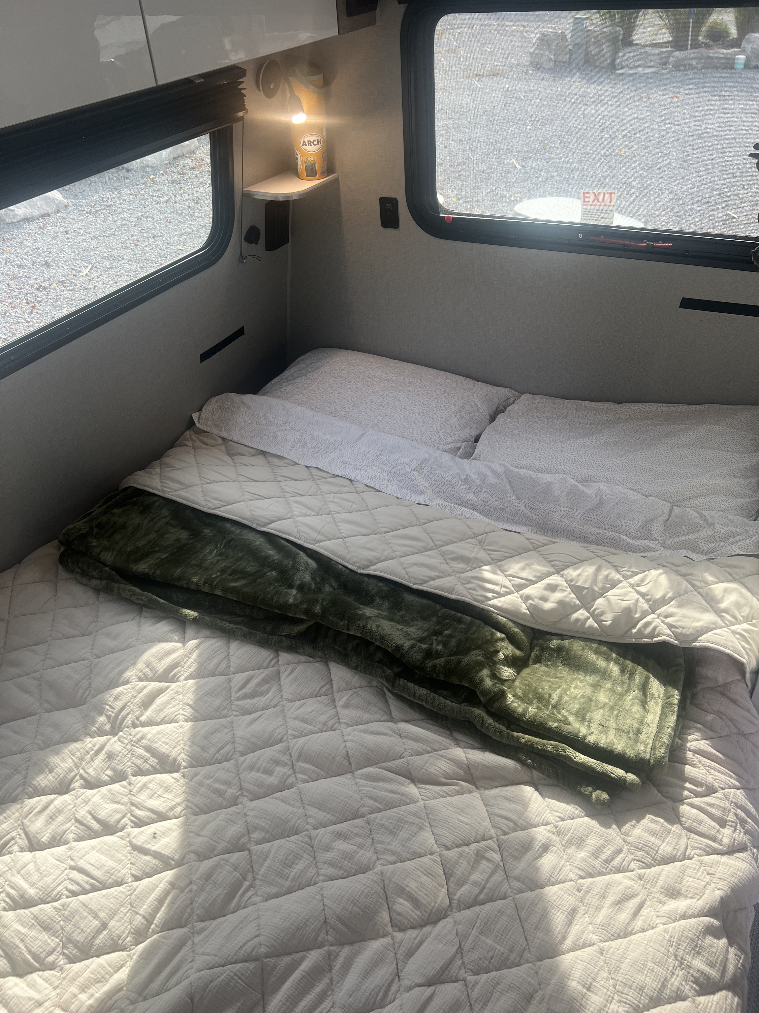 Relaxing RV rental interior. A made-up bed with pillows, a quilted comforter, and a green blanket, beside a sunlit window.. InTech RV Sol 2023