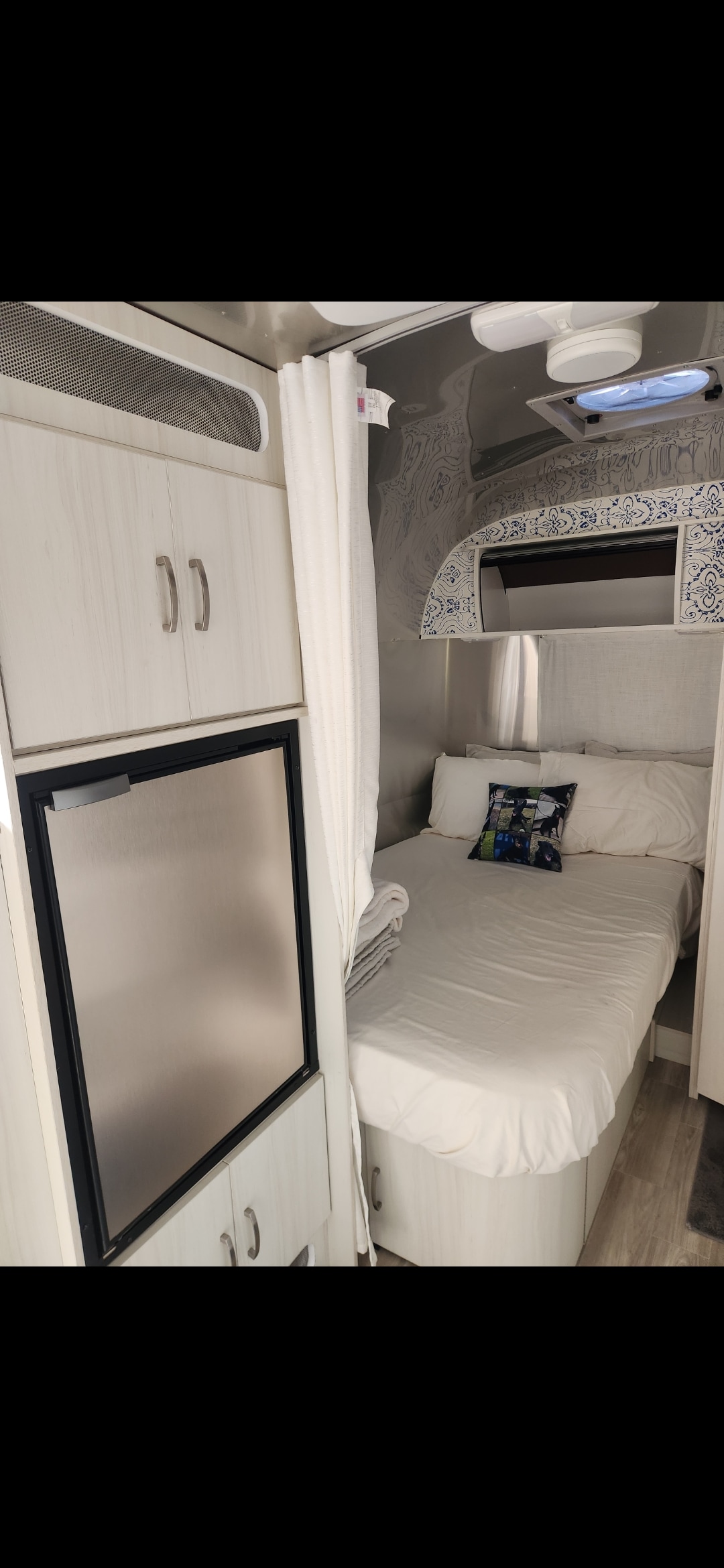 Bright rv rental interior features a cozy bed with white linens, a compact fridge, and light wood-grain cabinets.. Airstream Bambi 2023