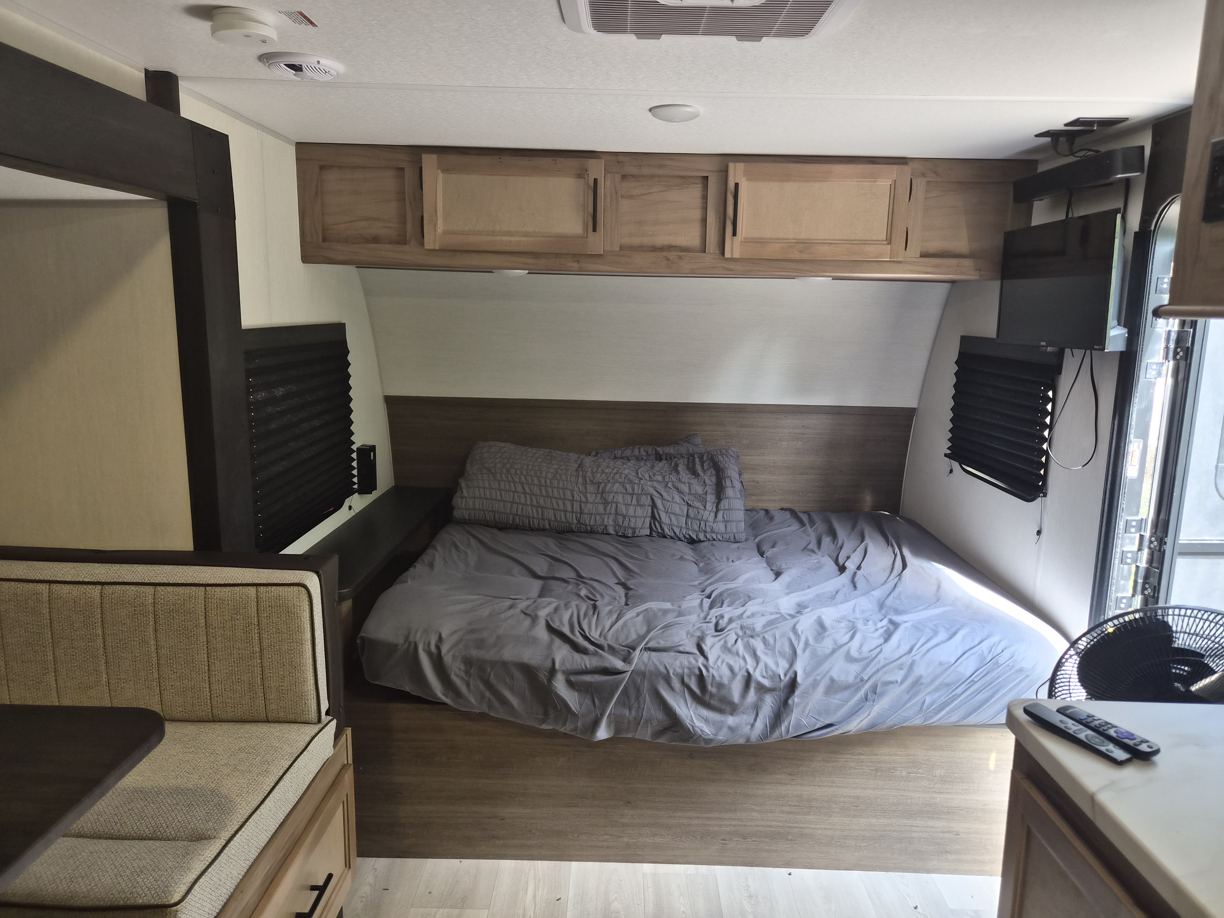This RV rental bedroom features a gray bed, overhead cabinets, wall-mounted TV, and a partial view of the dinette seating.. Forest River Cherokee Wolf Pup 2025