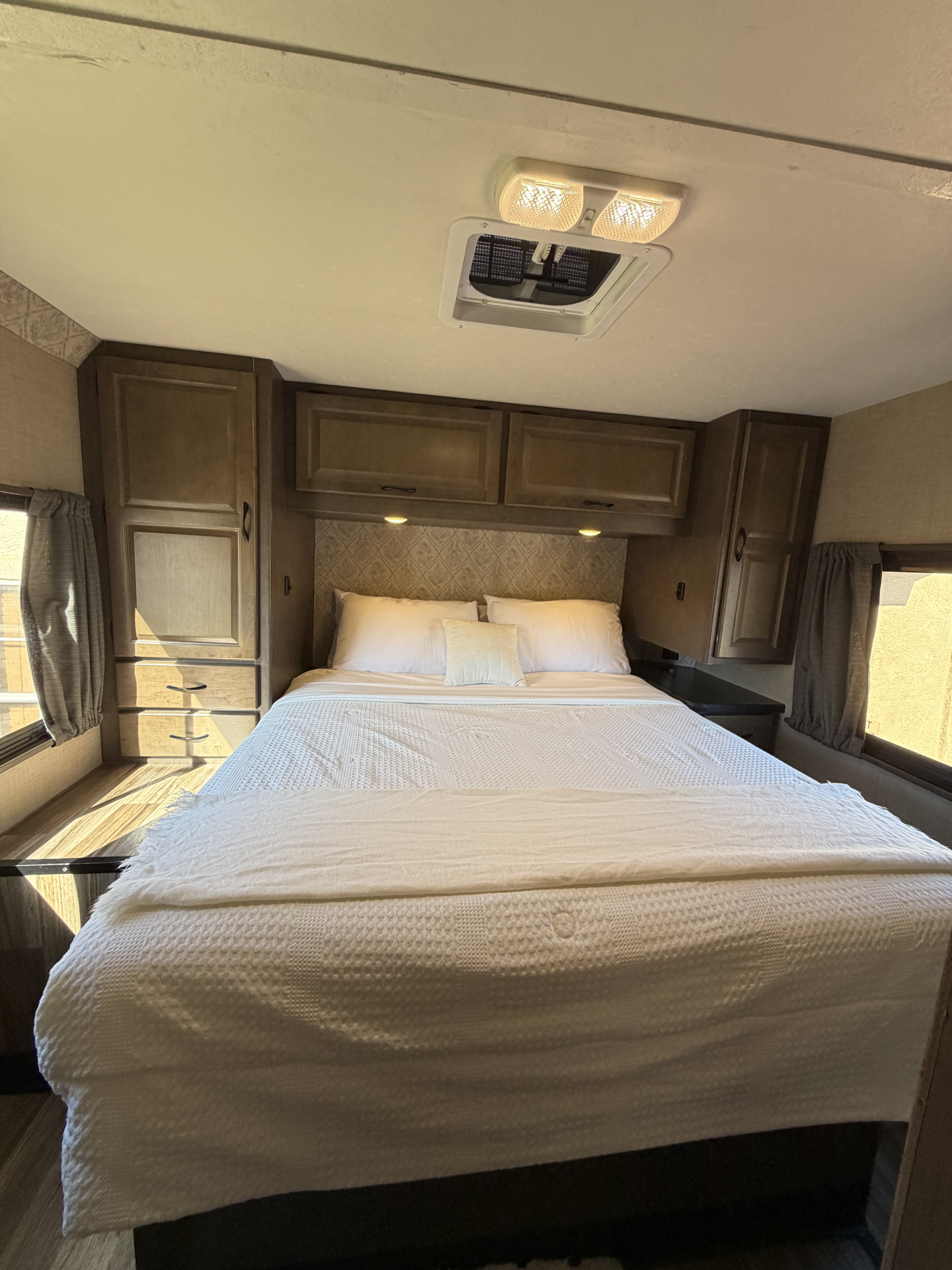 Cozy RV rental bedroom featuring a comfy bed with white linens, overhead cabinets, a ceiling light, and side windows.. Thor Majestic 2019