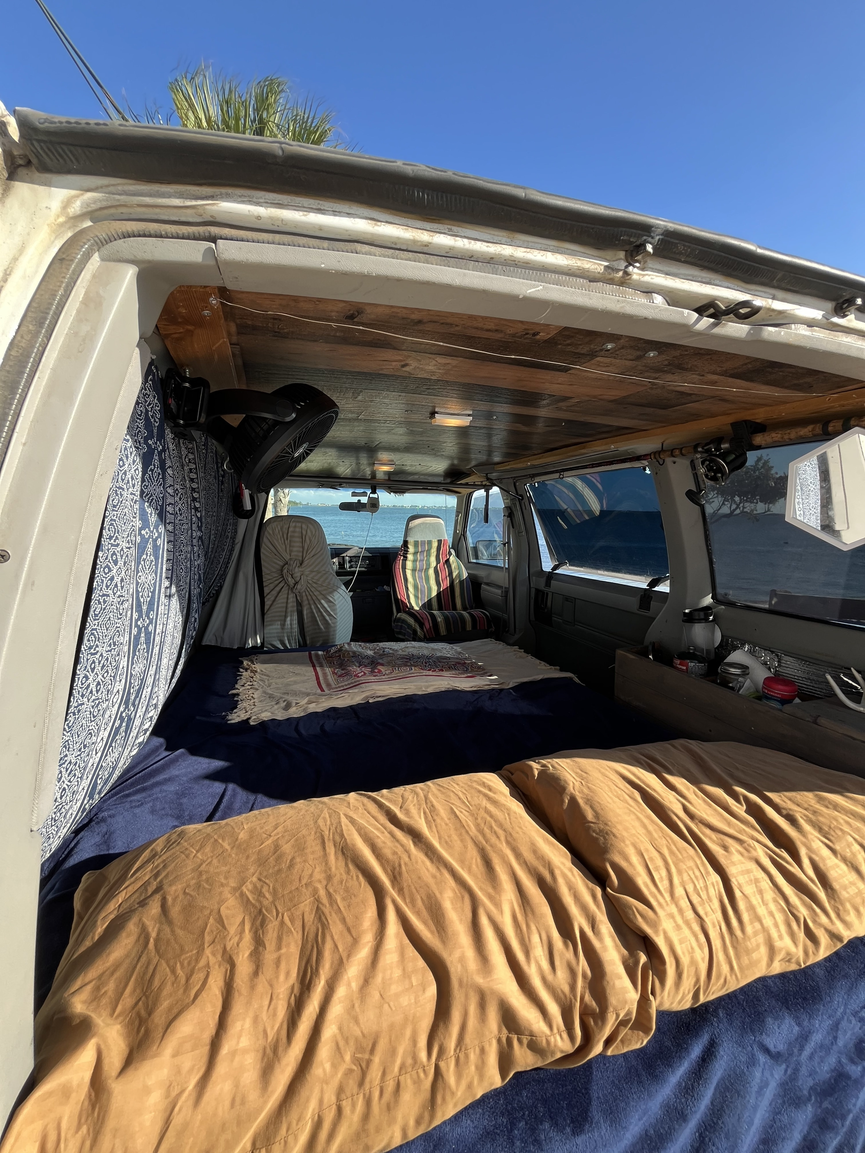 An RV rental's inviting interior: bed, custom wood ceiling, fan, and ocean view from a converted campervan.. Chevrolet Astro 1992