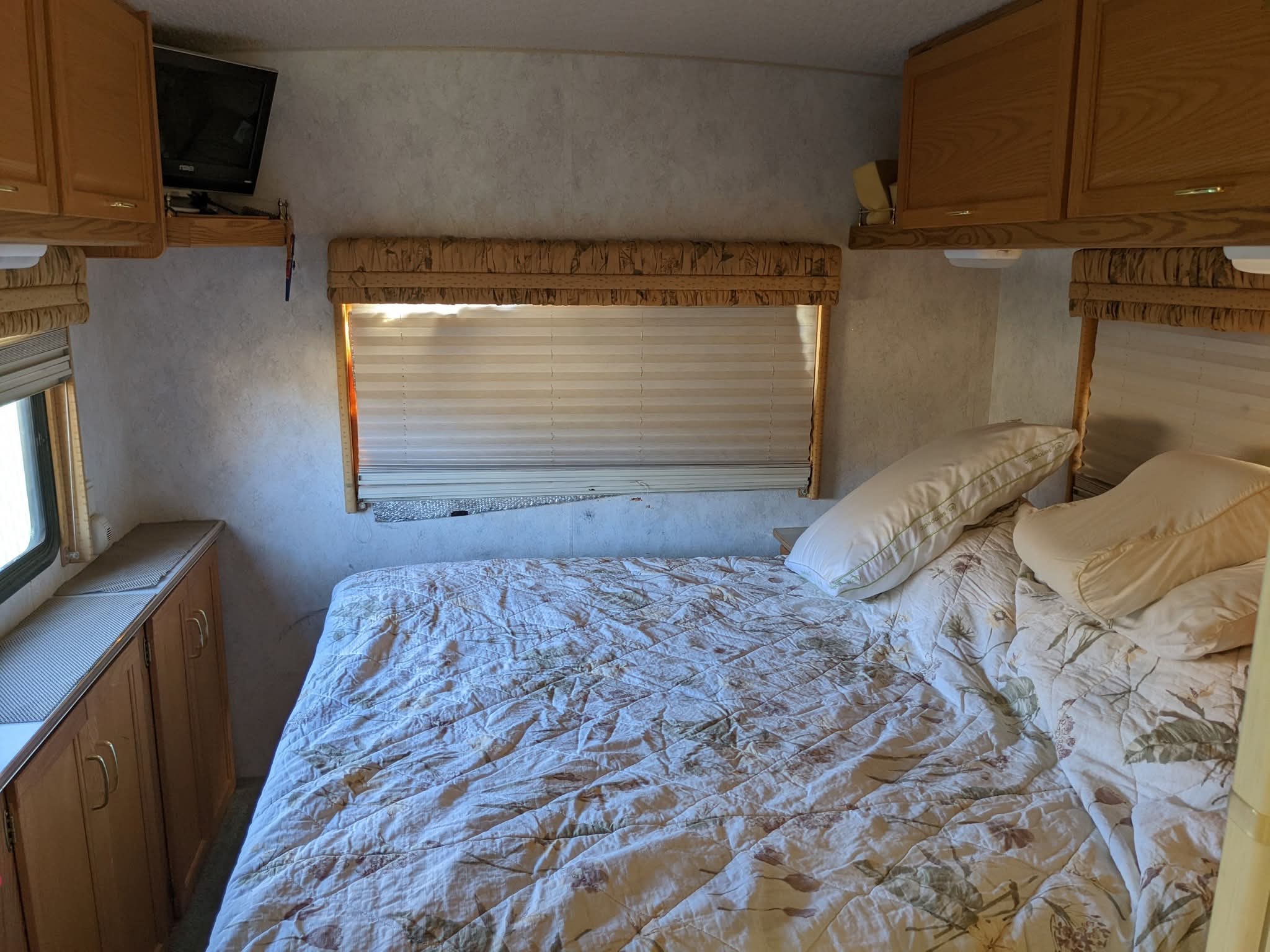 Cozy RV rental bedroom featuring a patterned comforter, pillows, overhead cabinets, a small TV, and window blinds.. Itasca Spirit 2003