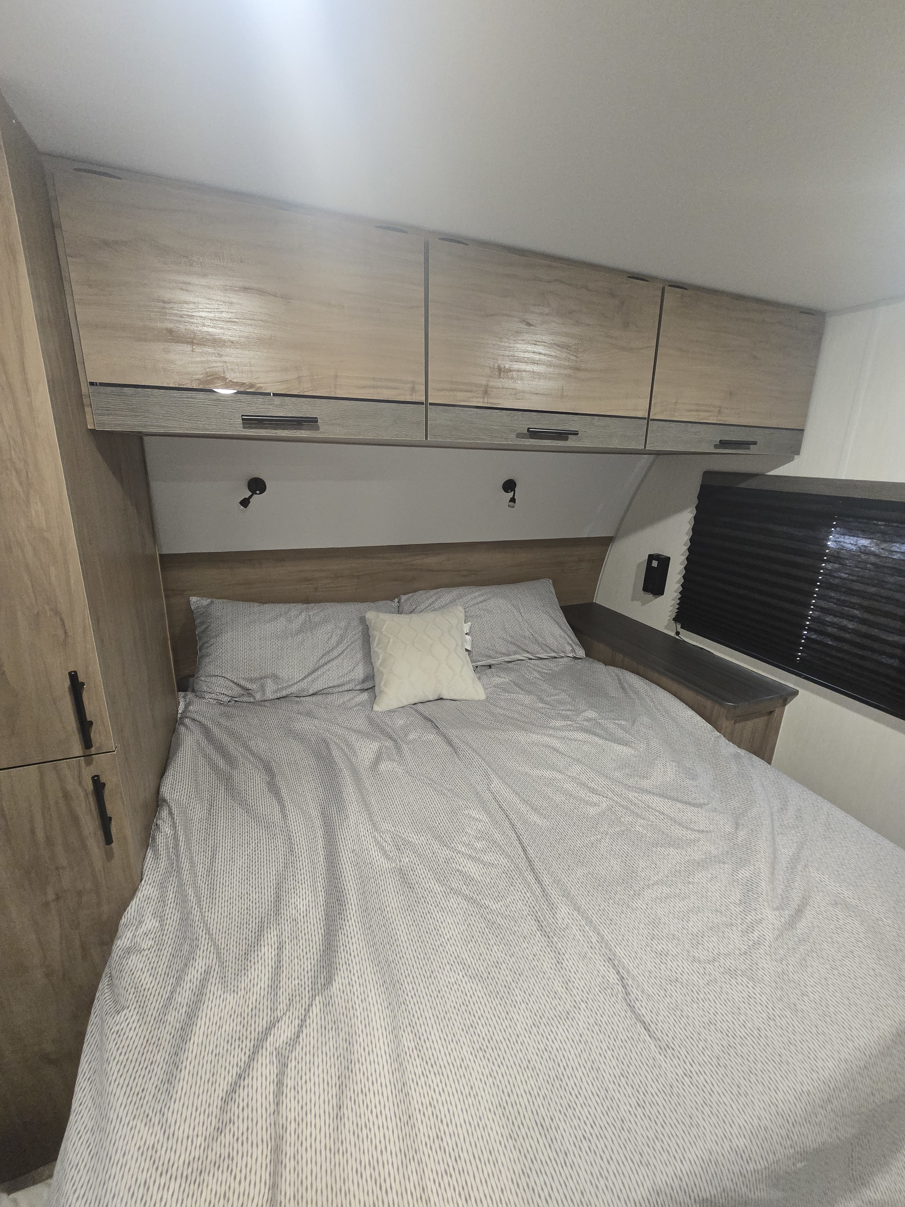 RV rental bedroom with light wood overhead storage, a comfortable striped bed, pillows, and bedside lighting.. Forest River Campsite Reserve 2025