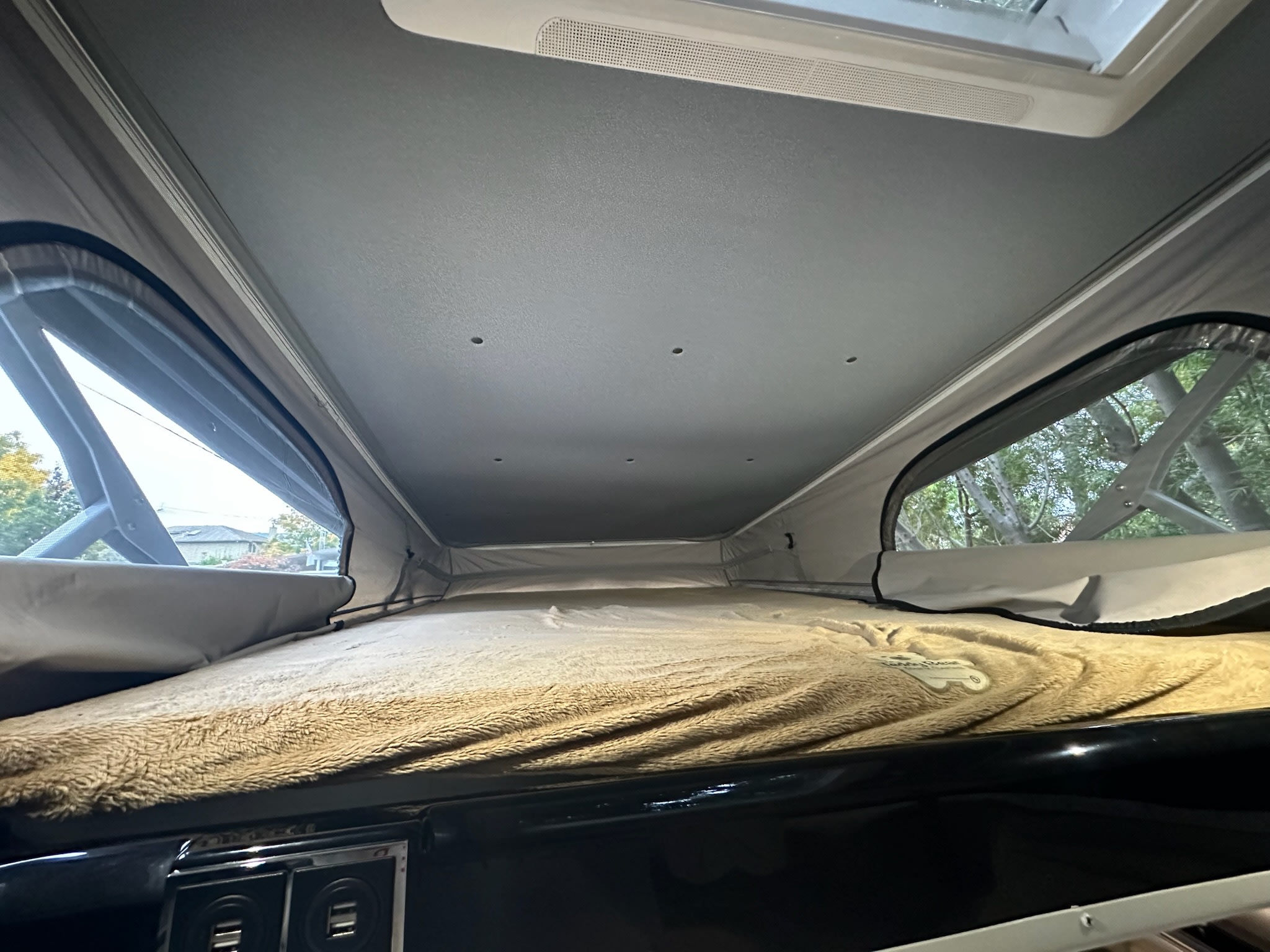 Cozy pop-top RV rental sleeping area with open windows showing trees, a comfortable fluffy bed, and convenient USB charging ports.. Thor Tellaro 2022