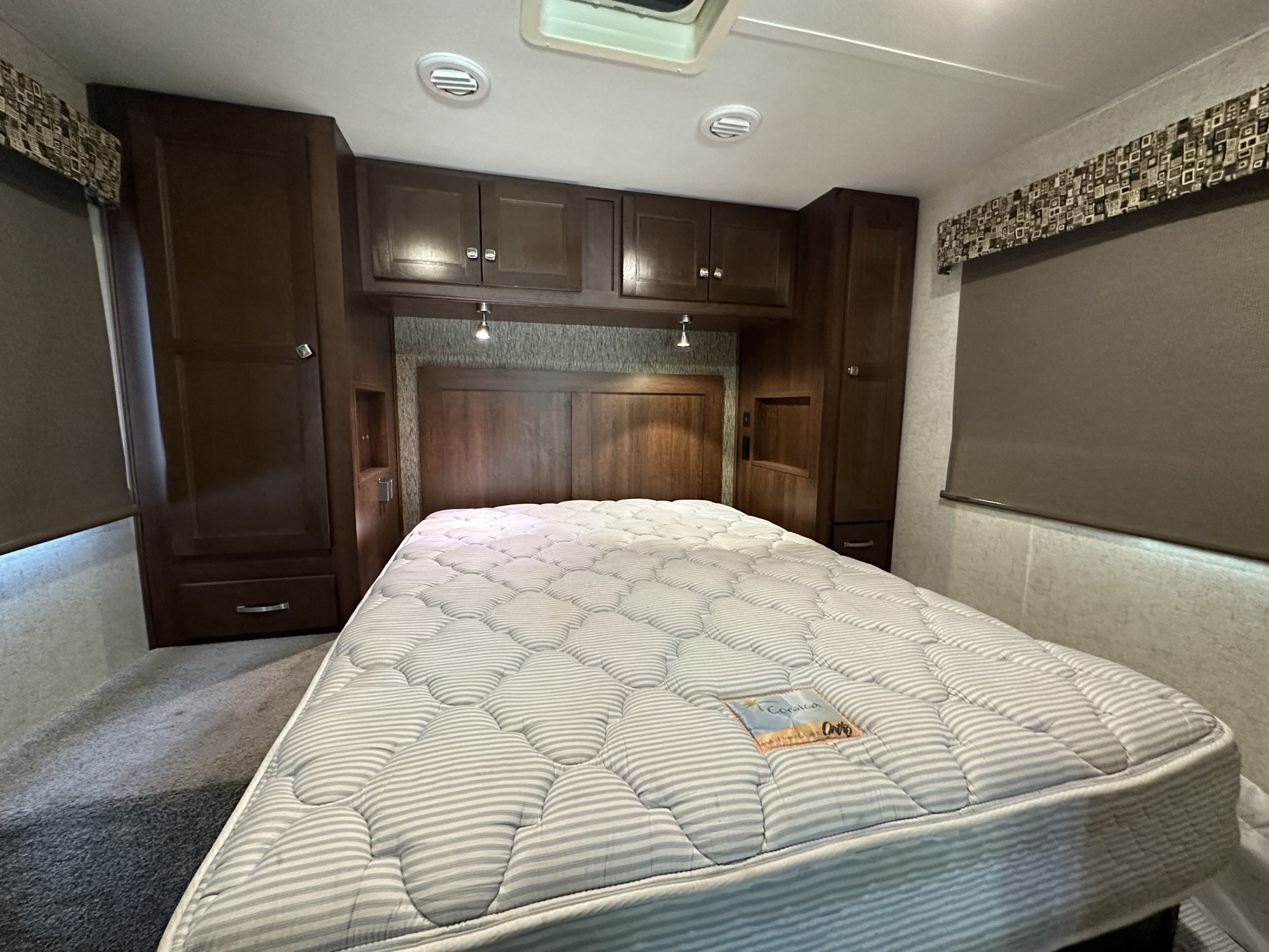 RV rental bedroom featuring a patterned mattress, dark wood cabinetry, overhead storage, and reading lights.. Forest River Sunseeker 2018