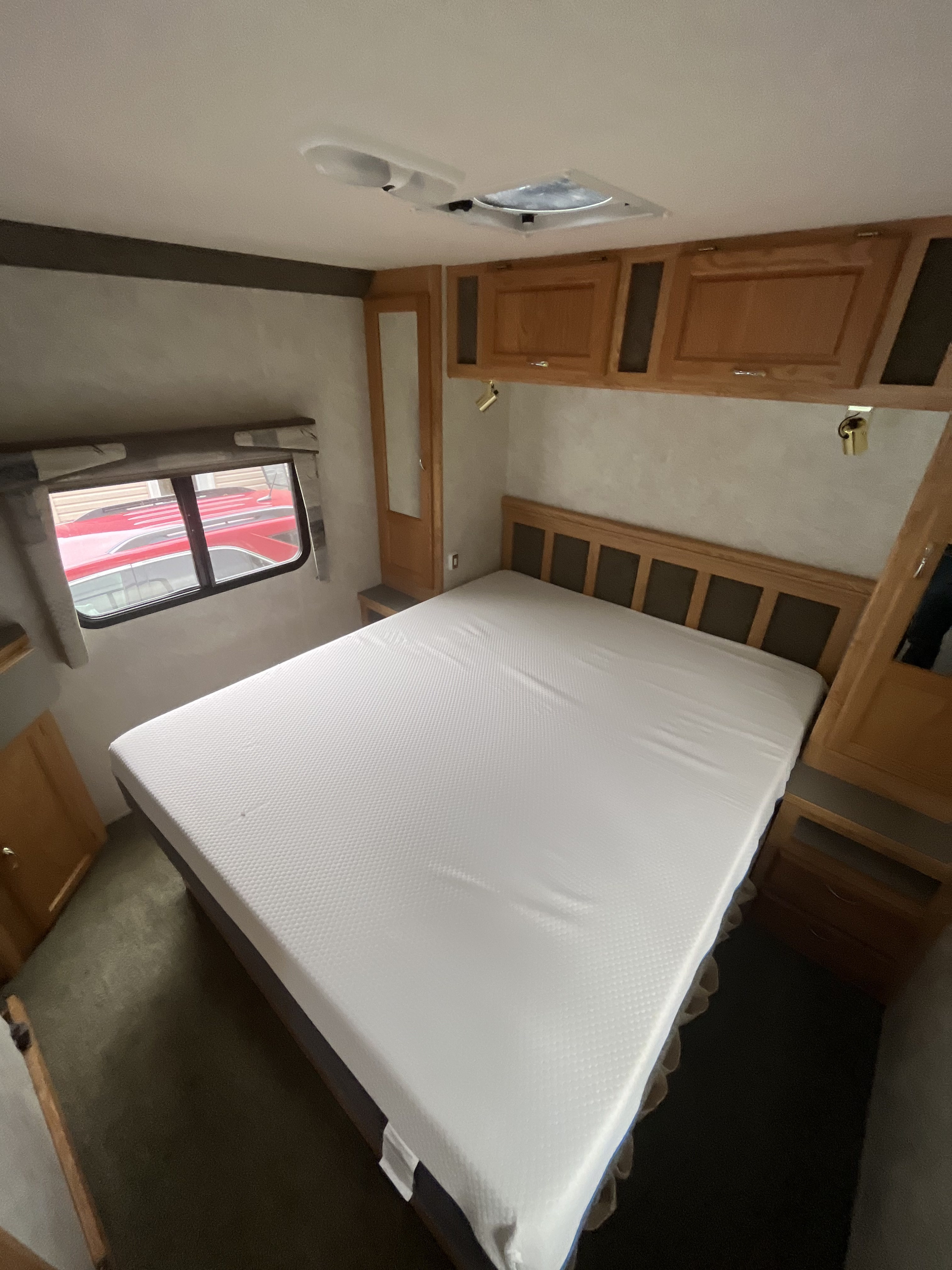 RV rental bedroom: a neat space with a bed, white mattress, wooden headboard, overhead cabinets, and a side window.. Bigfoot RV Bigfoot RV Motorhome 2004