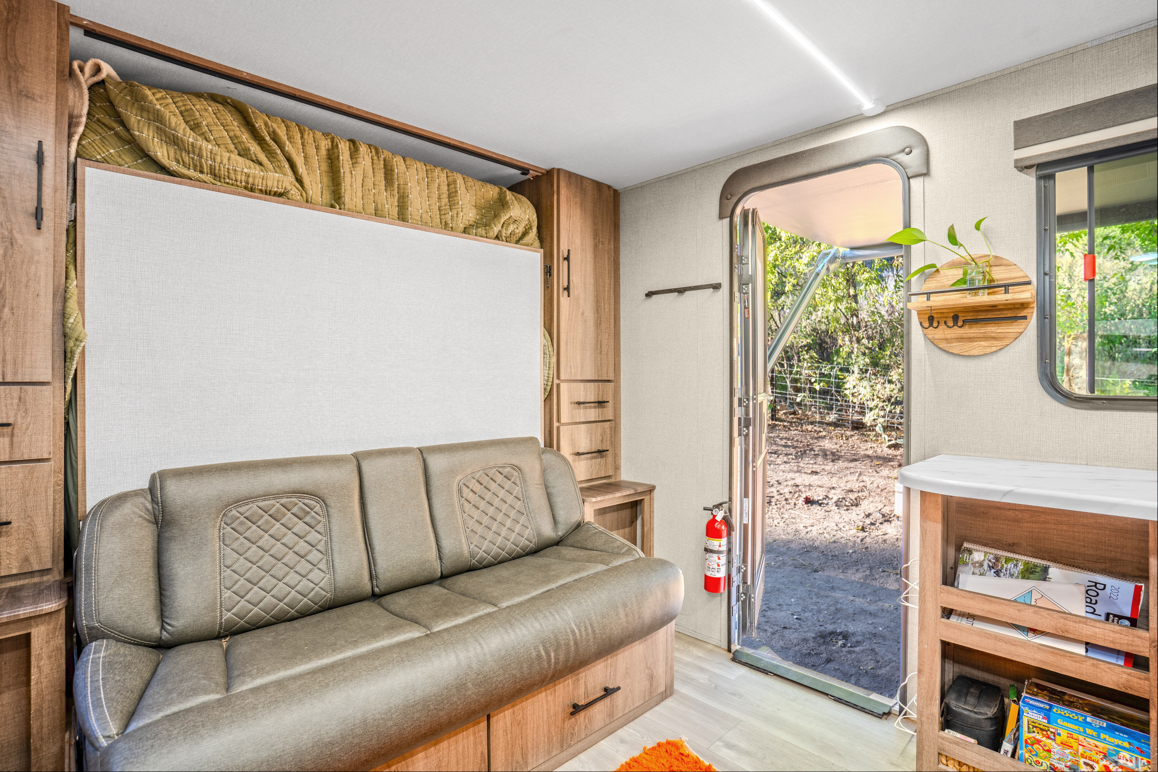 Compact rv rental living space with a pull-down bed over a sofa, wooden storage, and an open door inviting nature in.. Grand Design Imagine AIM 2024
