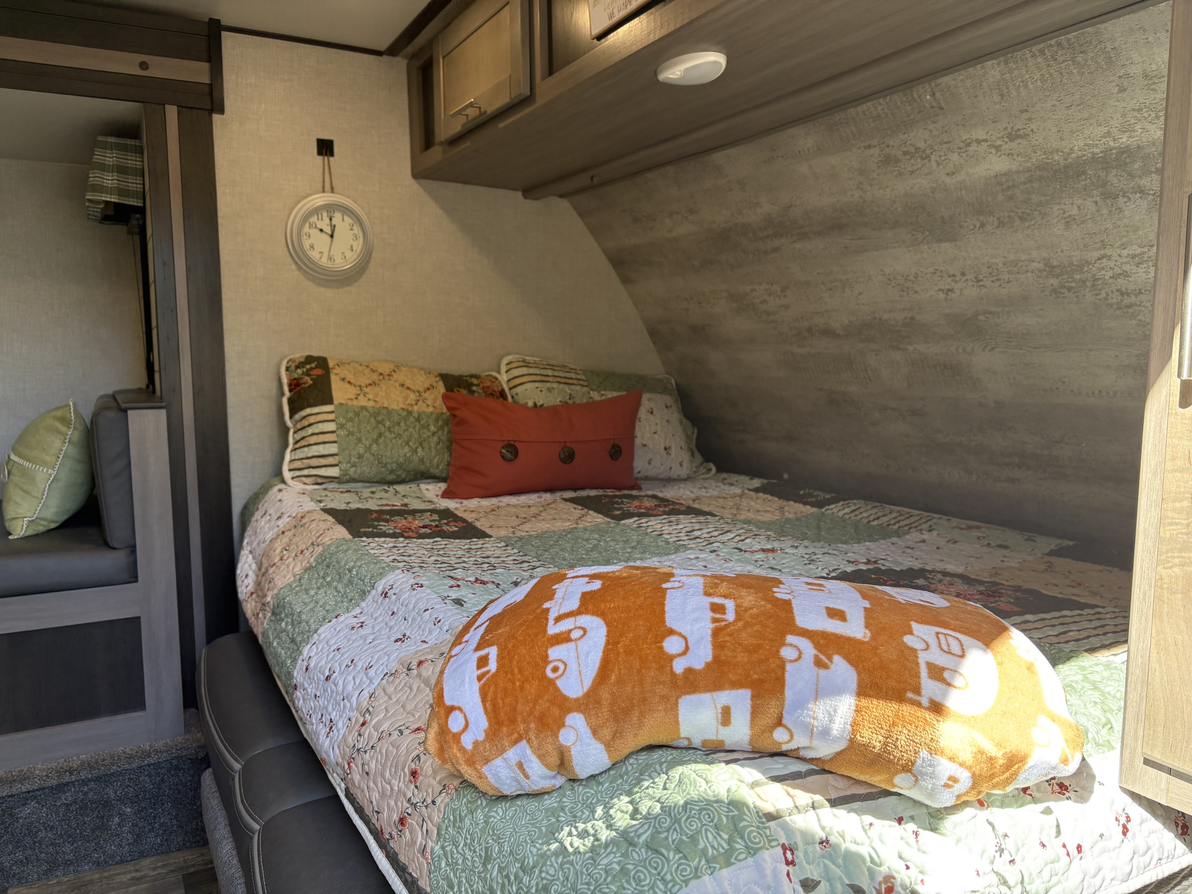 Cozy RV rental bedroom featuring a patchwork quilt, plush orange pillow with campers, and a wall clock.. Crossroads RV Sunset Trail Super Lite 2021