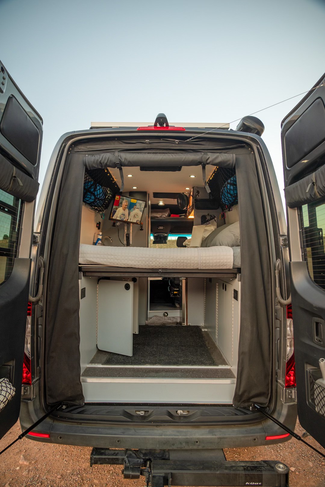 Spacious rv rental camper van interior with elevated bed, TV, and ample storage, viewed from the rear open doors.. Pleasure-Way Rekon 4x4 2022