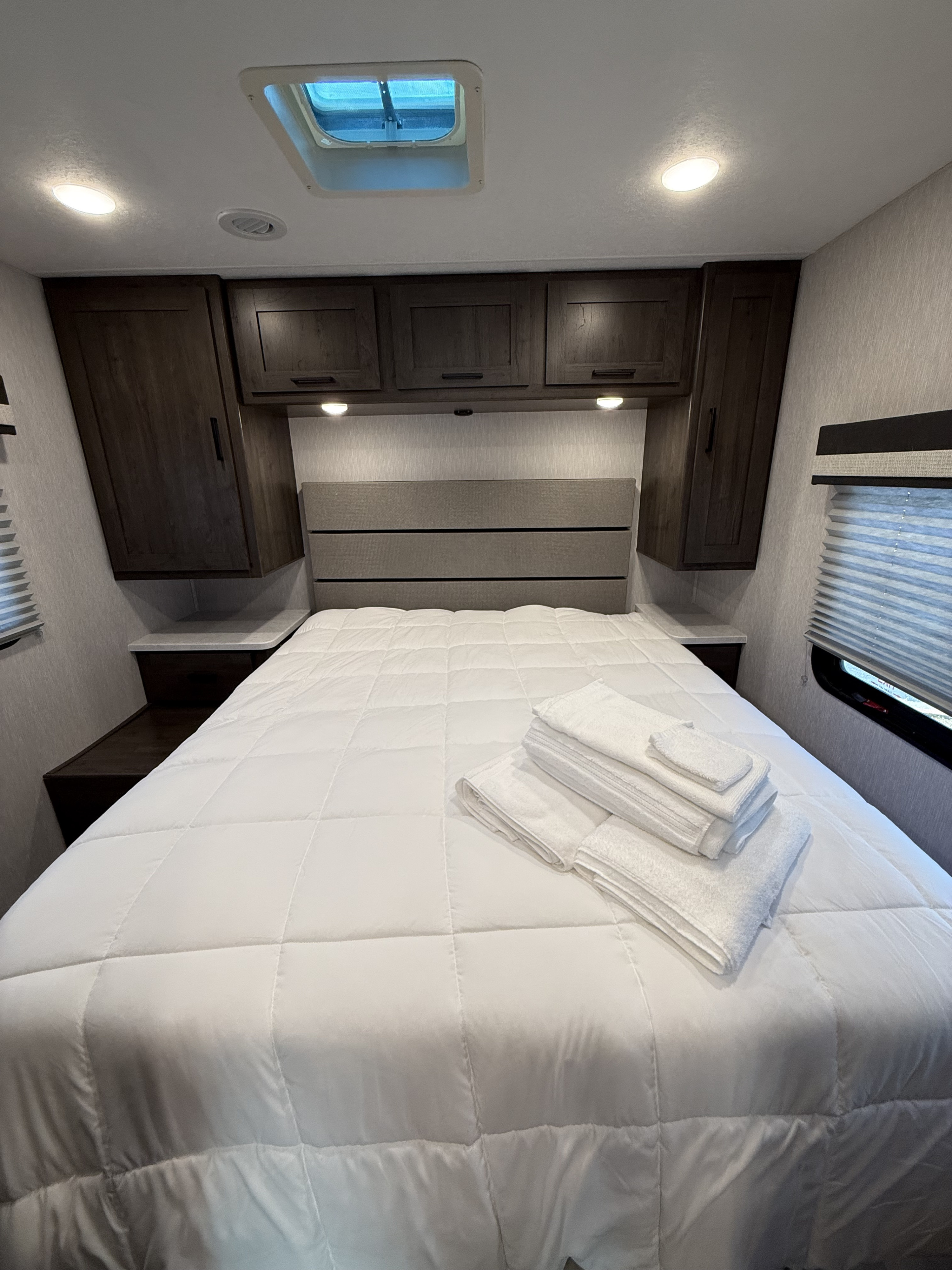 Cozy RV rental bedroom with a fresh white comforter, folded towels, dark wood cabinets, and a skylight for natural light.. Forest River Coachmen Pursuit 2024
