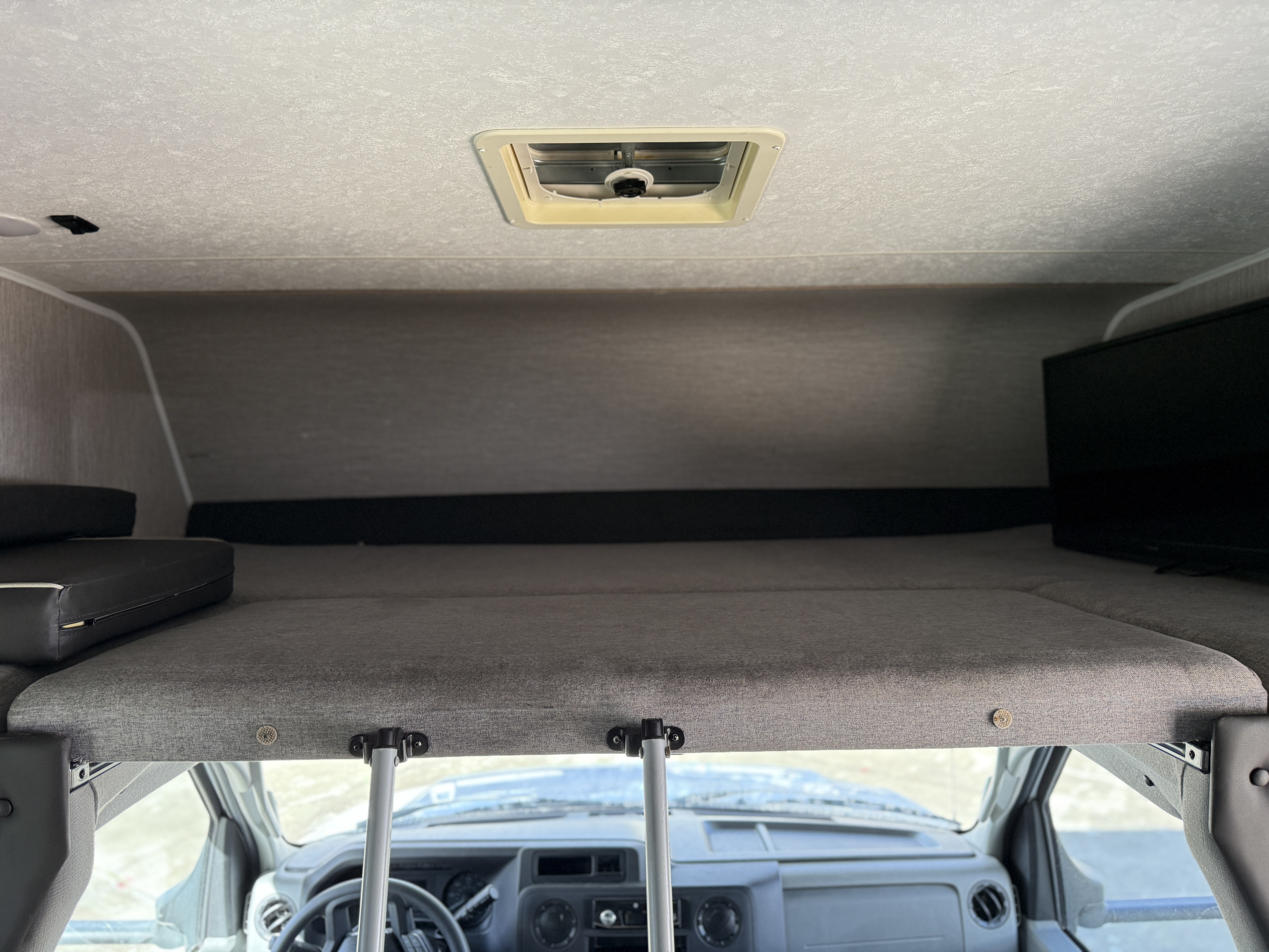 Overhead bunk in an RV rental, showing the gray mattress, a TV, and a roof vent above the driver's cab.. Coachmen Freelander 2021