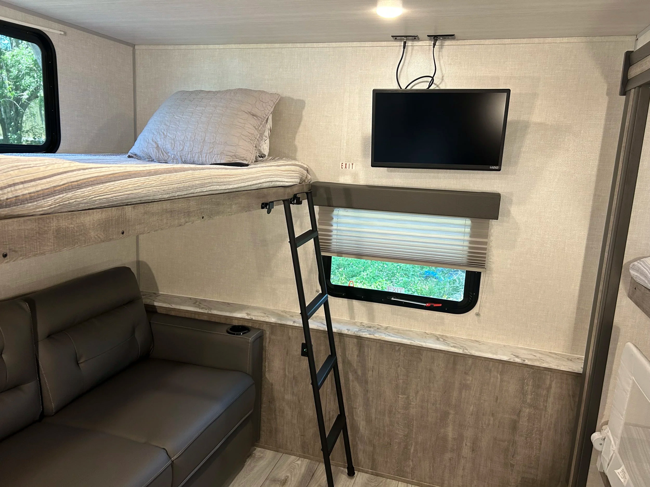 Inside an RV rental: a cozy bunk bed with a ladder, sofa below, wall-mounted TV, and window with outdoor views.. Forest River Surveyor 2023