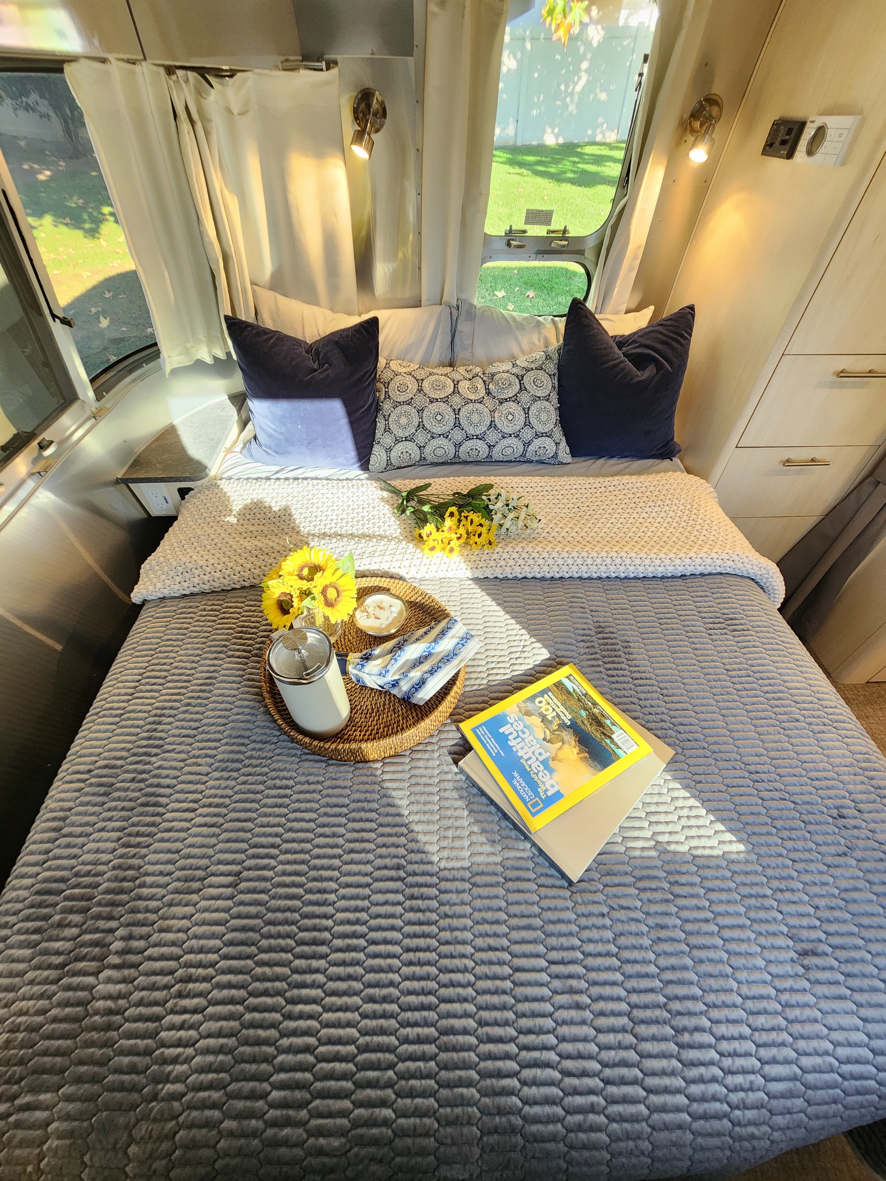 Cozy RV rental bedroom featuring a comfy bed, sunflowers, and reading materials, ready for your next adventure.. Airstream Flying Cloud 2022