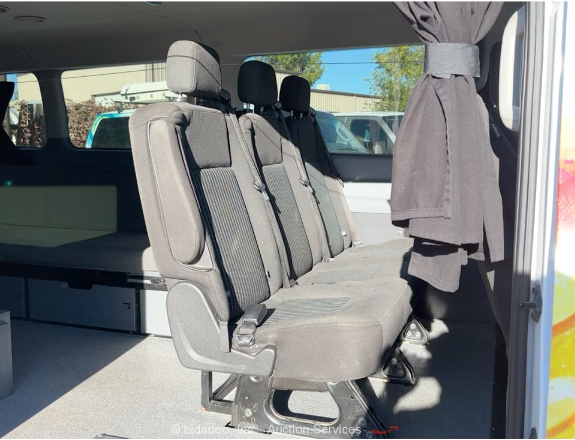 Inside an RV rental van: three black passenger seats, a bed/bench with storage drawers, and an open side door.. Ford Custom Transit 2017