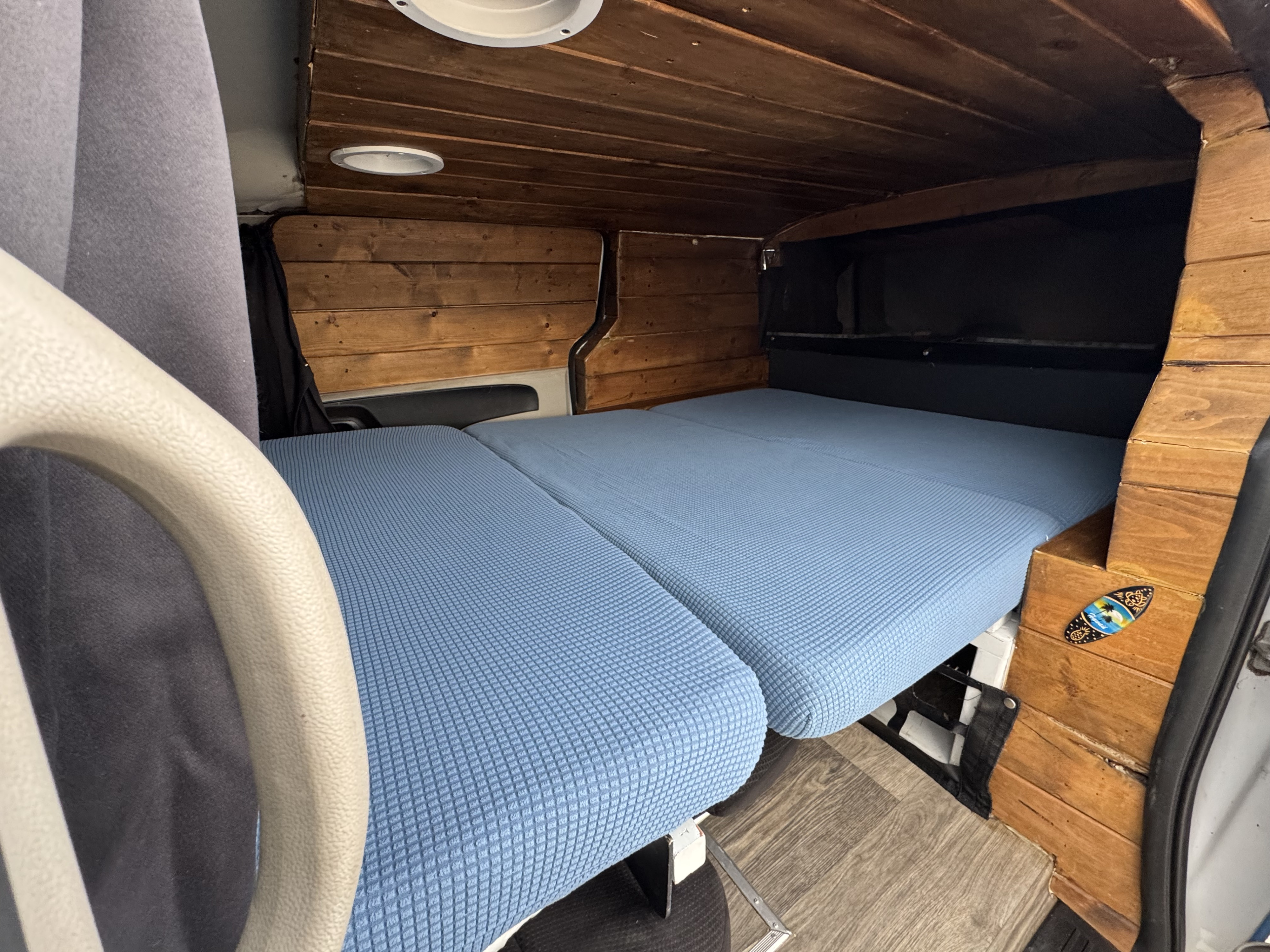 Compact rv rental interior with rustic wood paneling, featuring a blue modular bed and ceiling lights. Front handle visible.. Ram C/V Tradesman 2014
