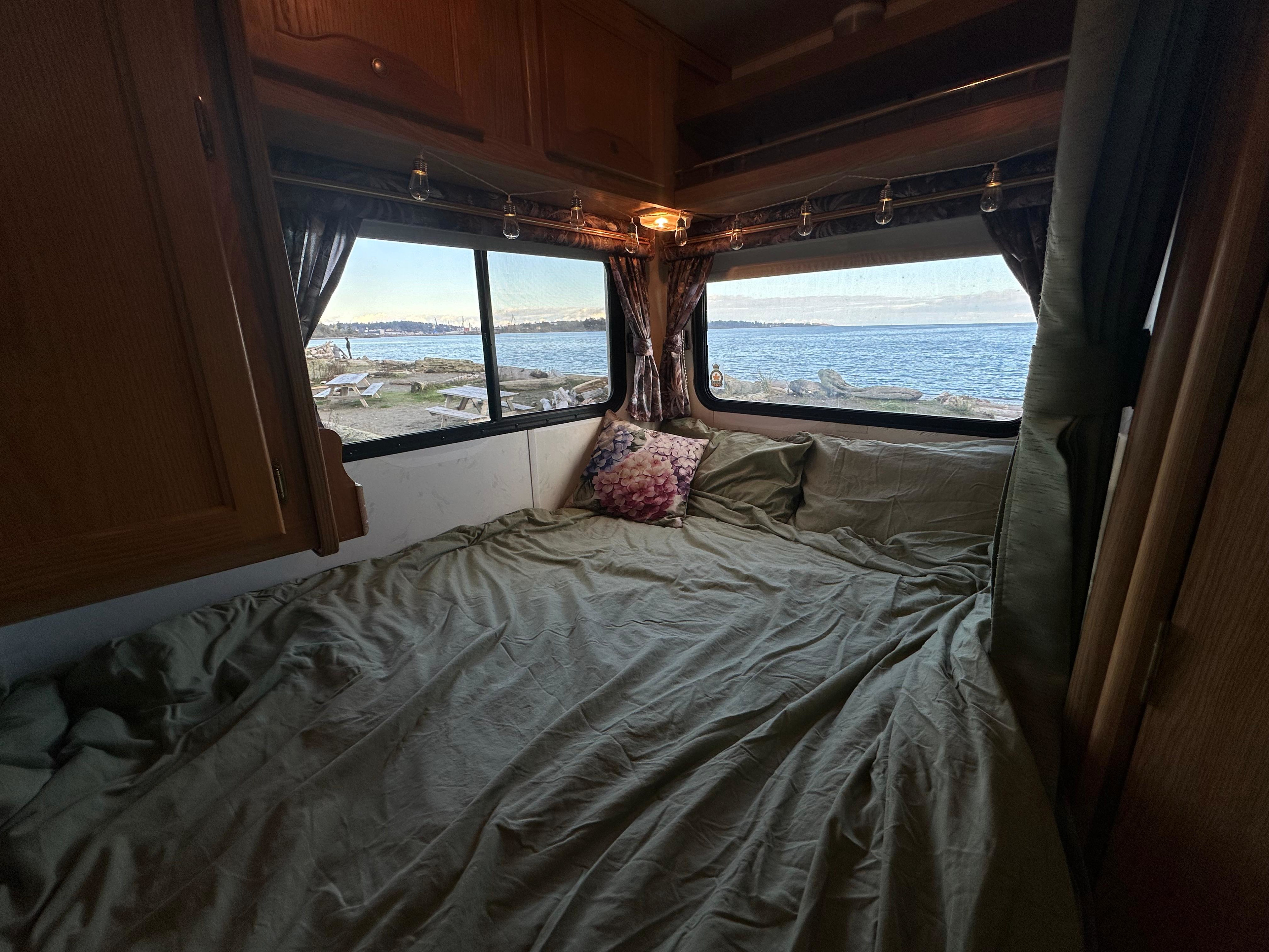 Cozy RV rental bedroom with an unmade bed, floral pillow, and panoramic windows revealing a calm ocean shore.. Ford E350 Travelmaster 1997