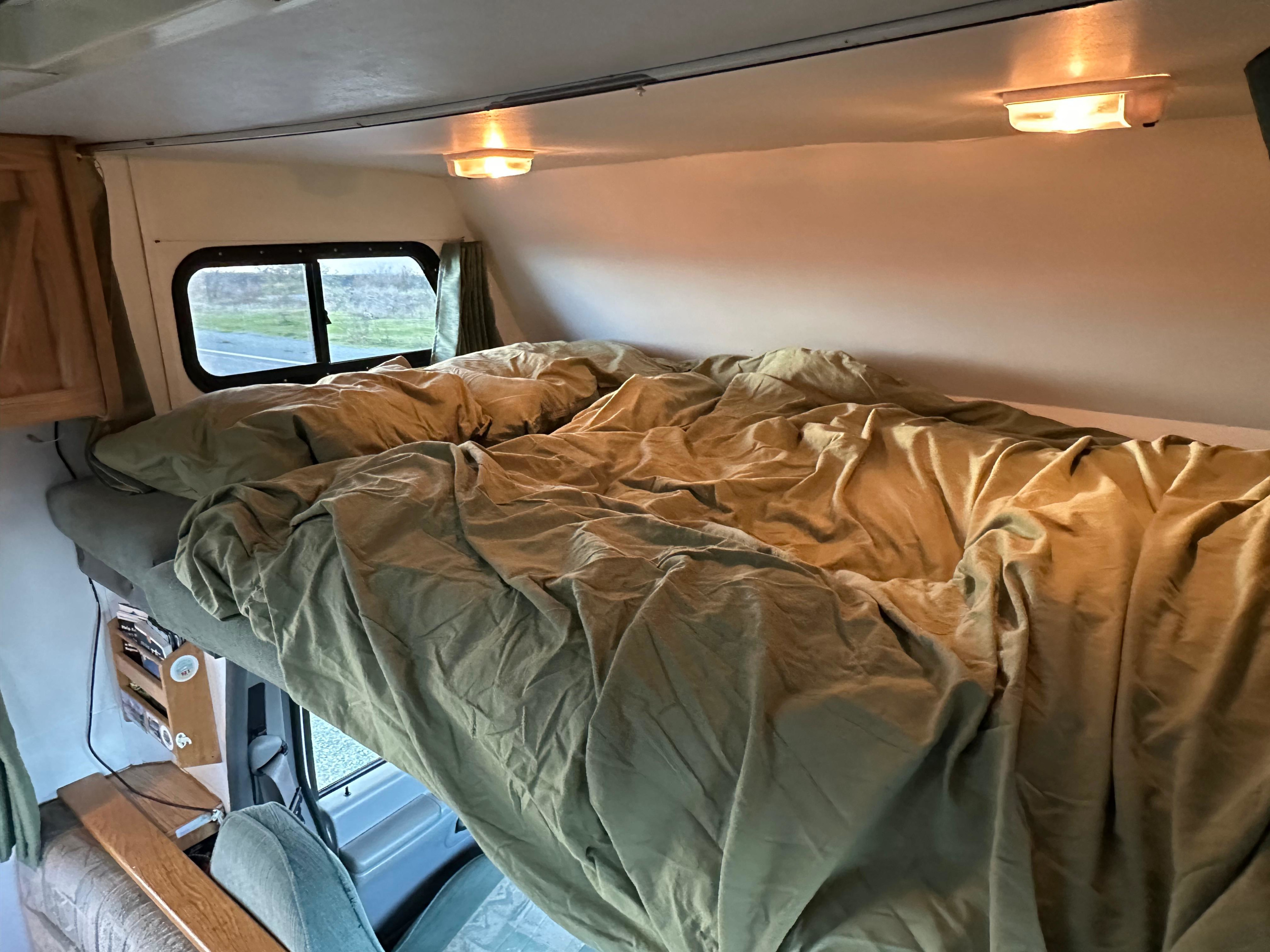 Cozy rv rental overhead bed with olive bedding and warm lighting, offering window views. Ideal for comfortable travel.. Ford E350 Travelmaster 1997