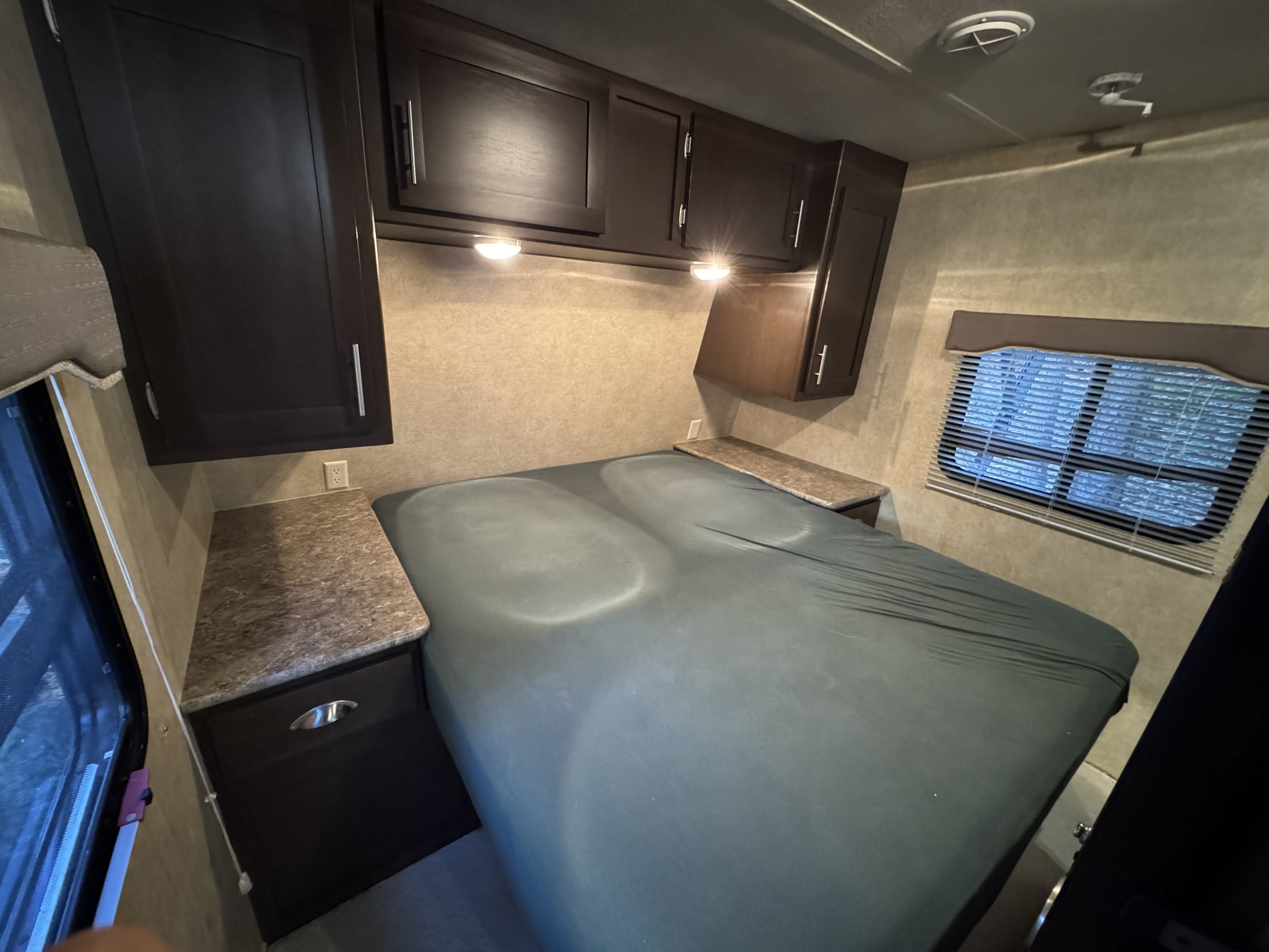 An RV rental bedroom with a bed, dark wood cabinets, side counters with outlets, and windows with blinds for a compact interior.. Forest River EVO 2013