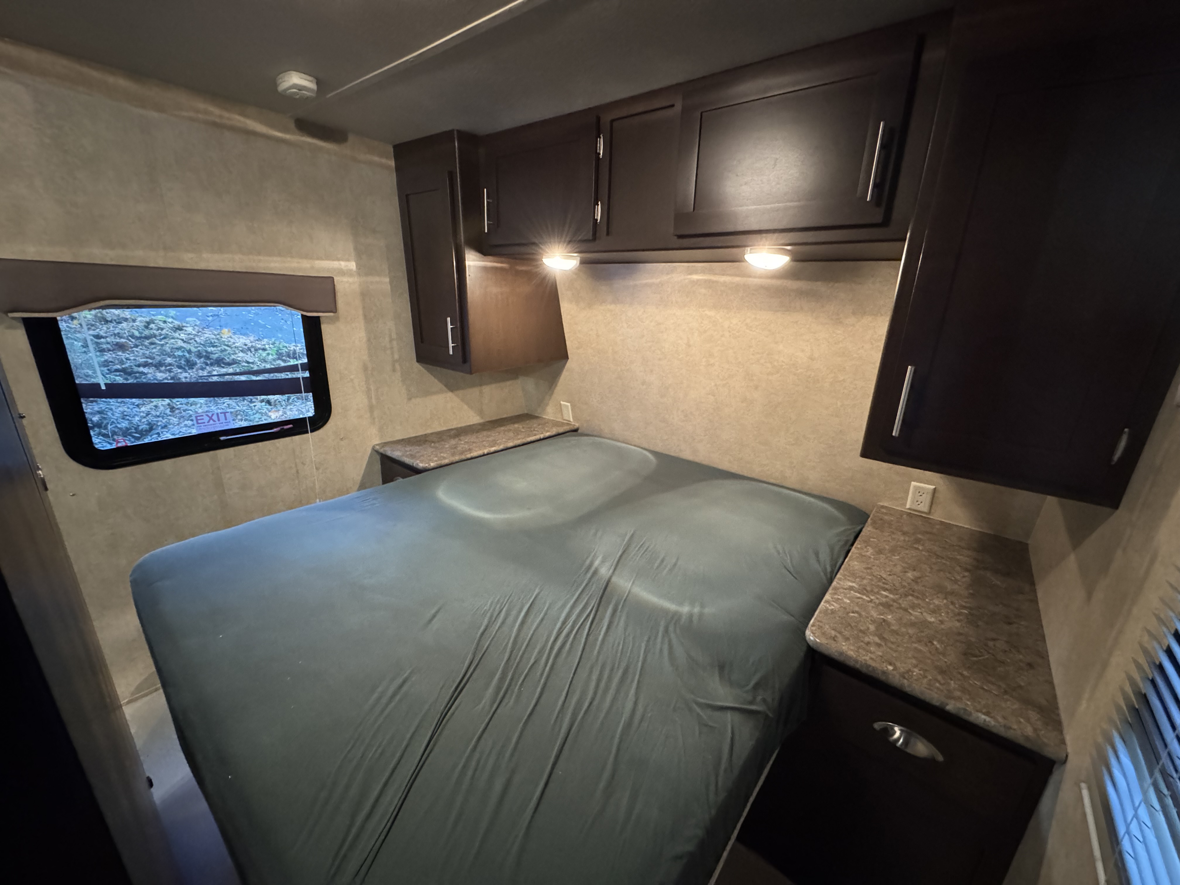 Cozy RV rental bedroom with a comfy bed, overhead dark wood cabinets, bedside tables, and a window for scenic views.. Forest River EVO 2013