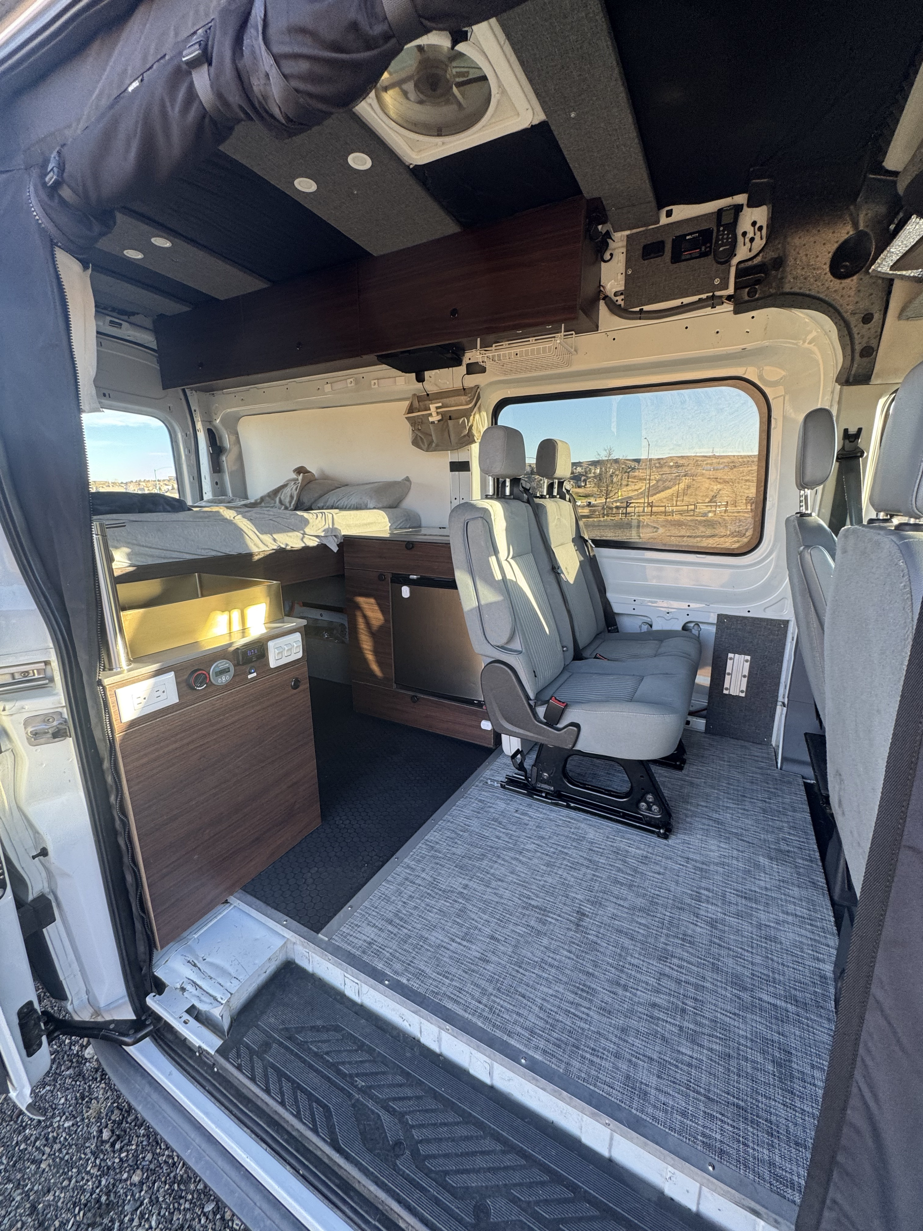 Inside this RV rental camper van, a bed, kitchen, and passenger seats are visible from the open side door.. Ford Transit 2018