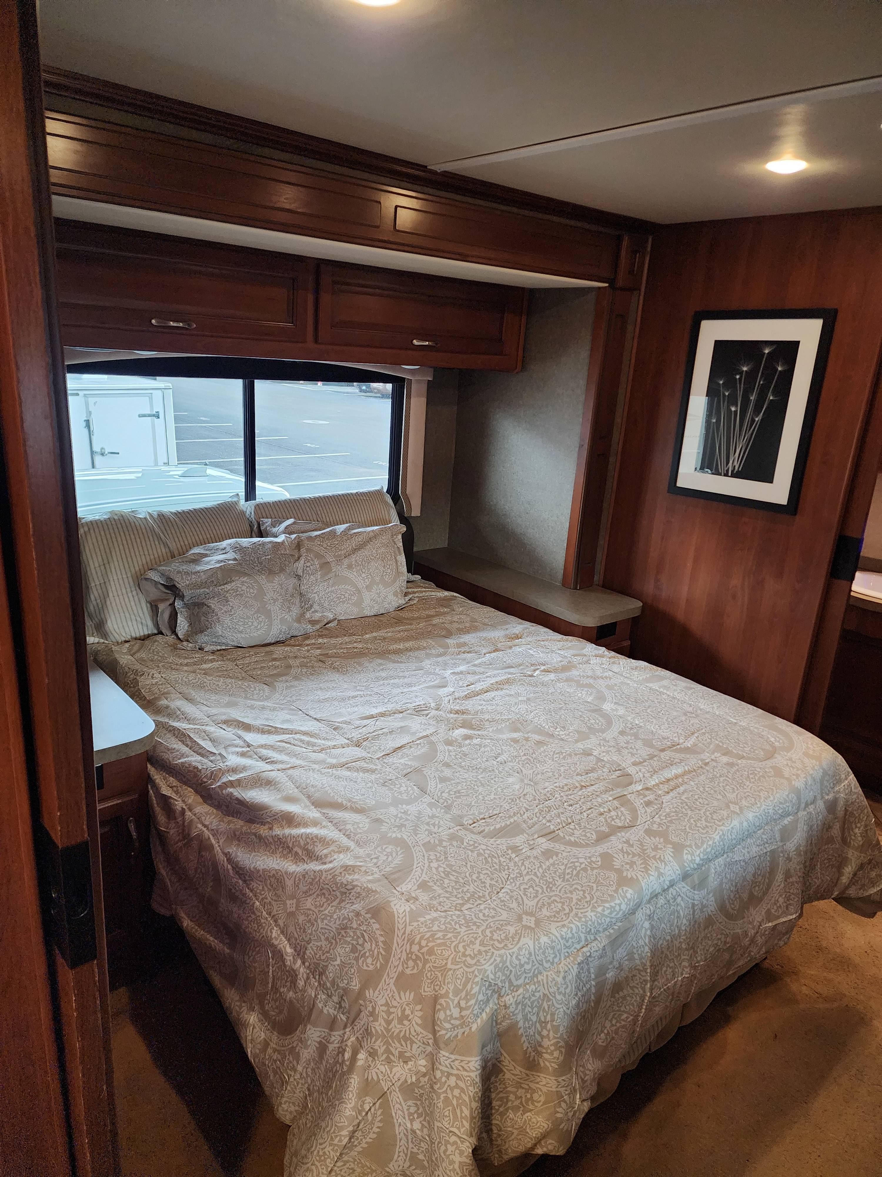 Cozy RV rental bedroom featuring a queen-sized bed with patterned bedding, overhead wood cabinets, and a side window.. Fleetwood Bounder 35k 2016