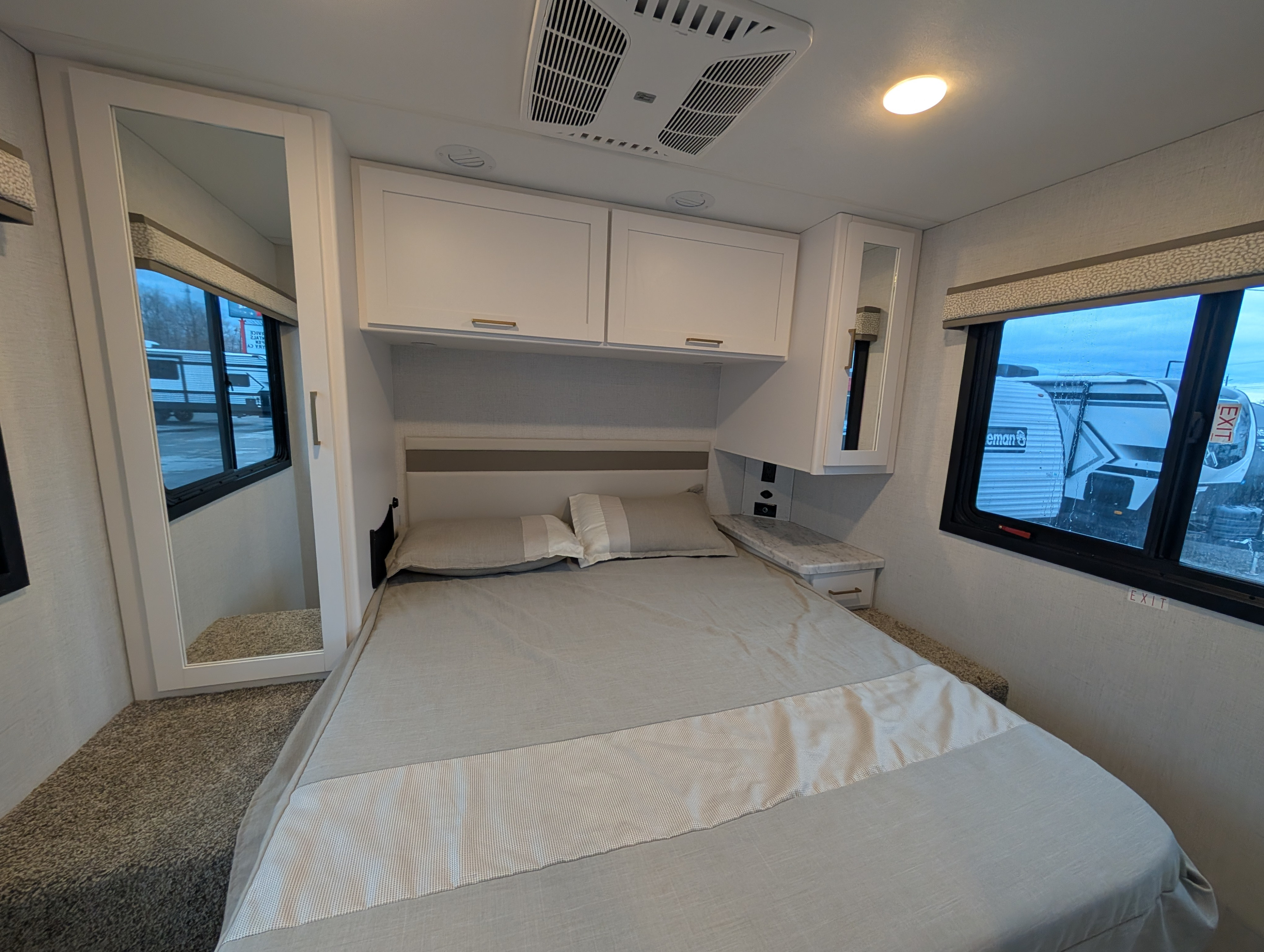 A cozy RV rental bedroom features a gray bed, white overhead cabinets, side mirrors, and large windows with outdoor views.. THOR CHATEAU 2026